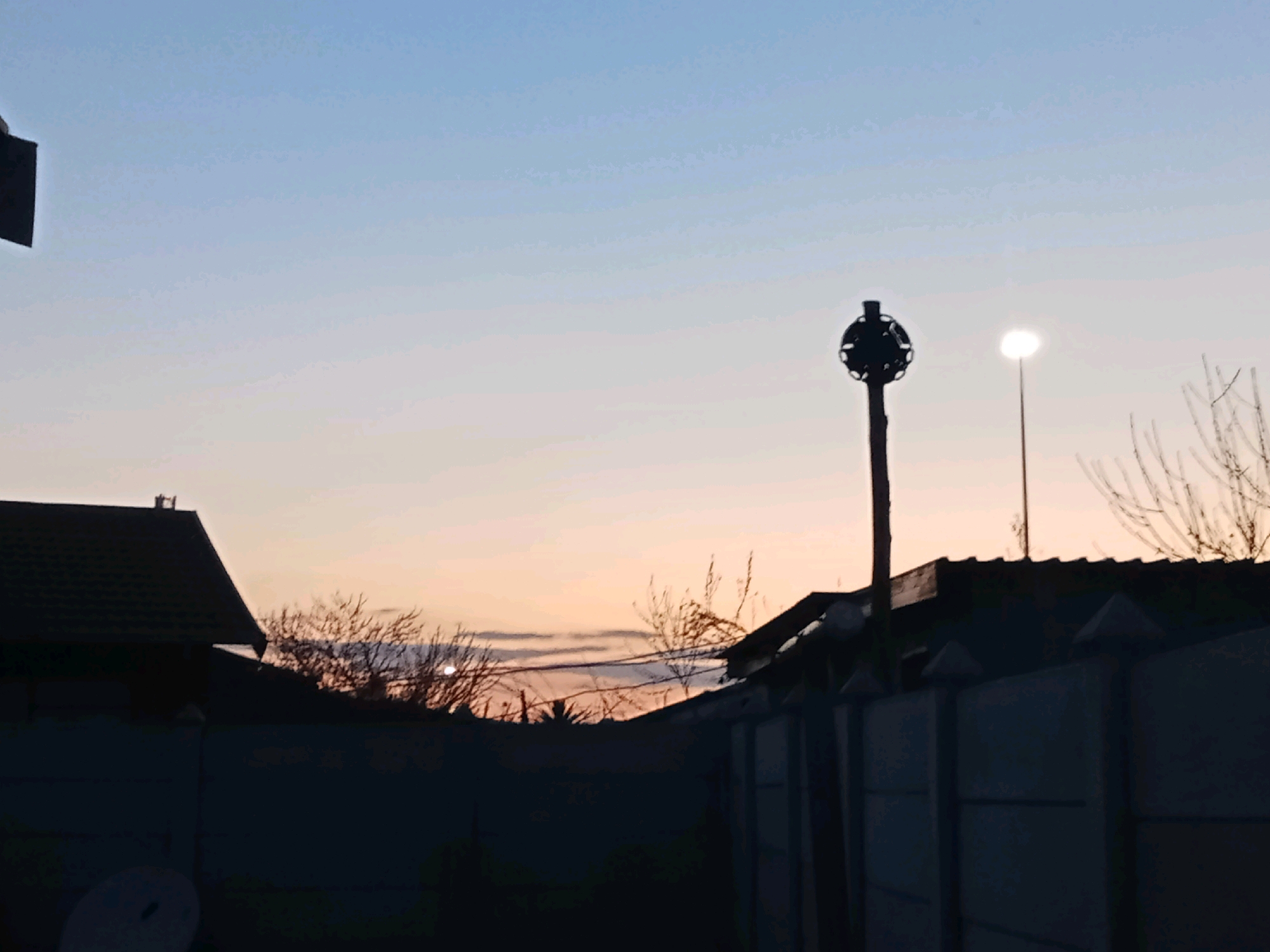 The image captures a twilight scene in Meloding, South Africa. The sky displays a gradient of blues and oranges, indicative of either dawn or dusk. In the foreground, silhouetted against the sky, is a concrete fence topped with pointed concrete caps. To the left, the dark silhouette of a house with a tiled roof is visible. A satellite dish is also faintly discernible in the lower left corner.
Dominating the mid-ground is a tall pole supporting a circular, cage-like structure with various protrusions, resembling a siren or some form of communication equipment. Further to the right, a street lamp with its light illuminated stands tall. In the background, bare-branched trees are silhouetted against the colorful sky. The overall atmosphere is serene and quiet, typical of a residential area at the transition between day and night.