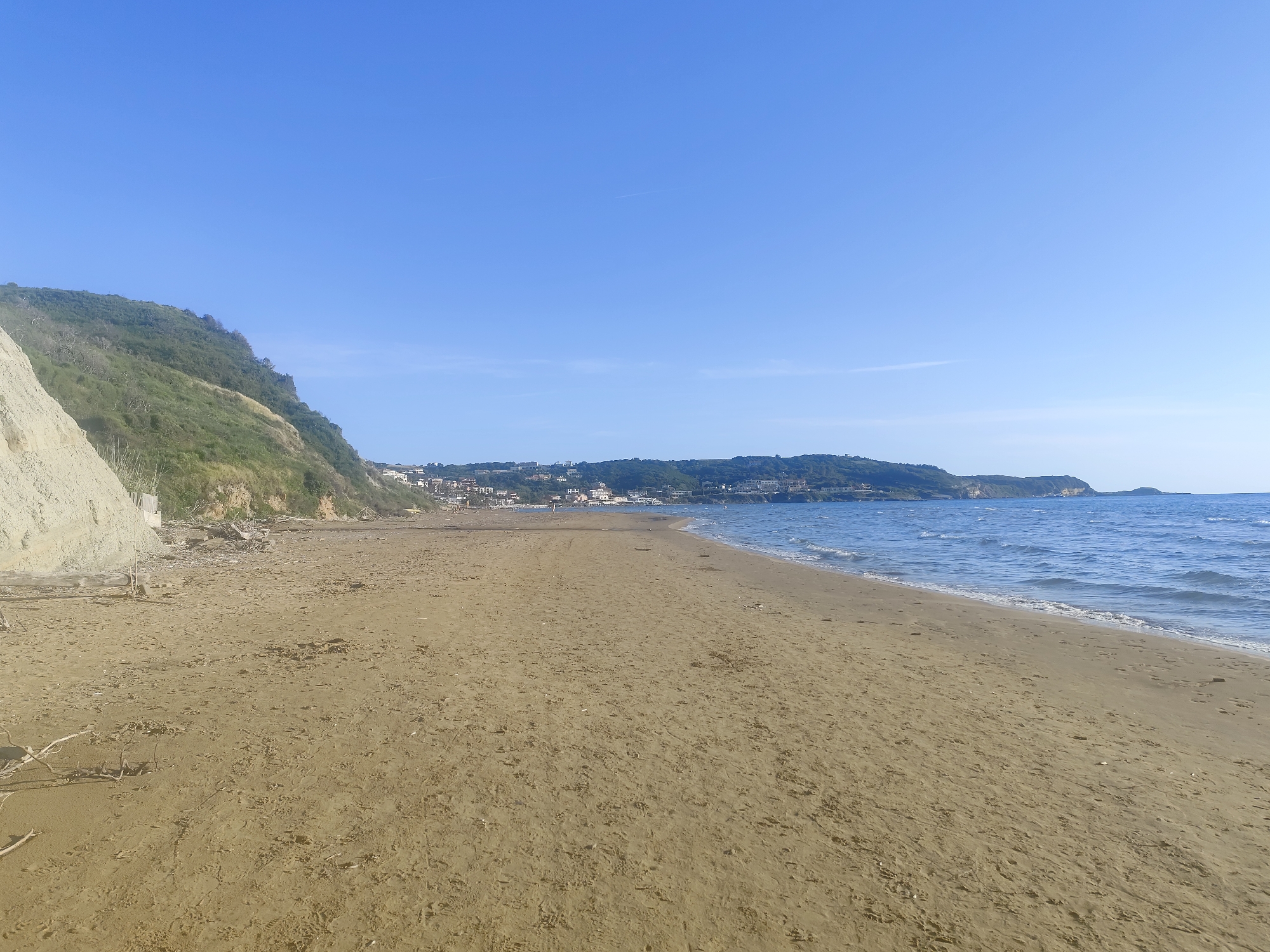 This image depicts a sandy beach on a sunny day in an unknown location. The sky is clear blue. To the left, a high cliff rises from the shoreline. Along the coastline, there's vegetation and some fallen debris. In the distance, houses and buildings are visible. The ocean waves gently wash the sandy shore. The beach is otherwise devoid of people.