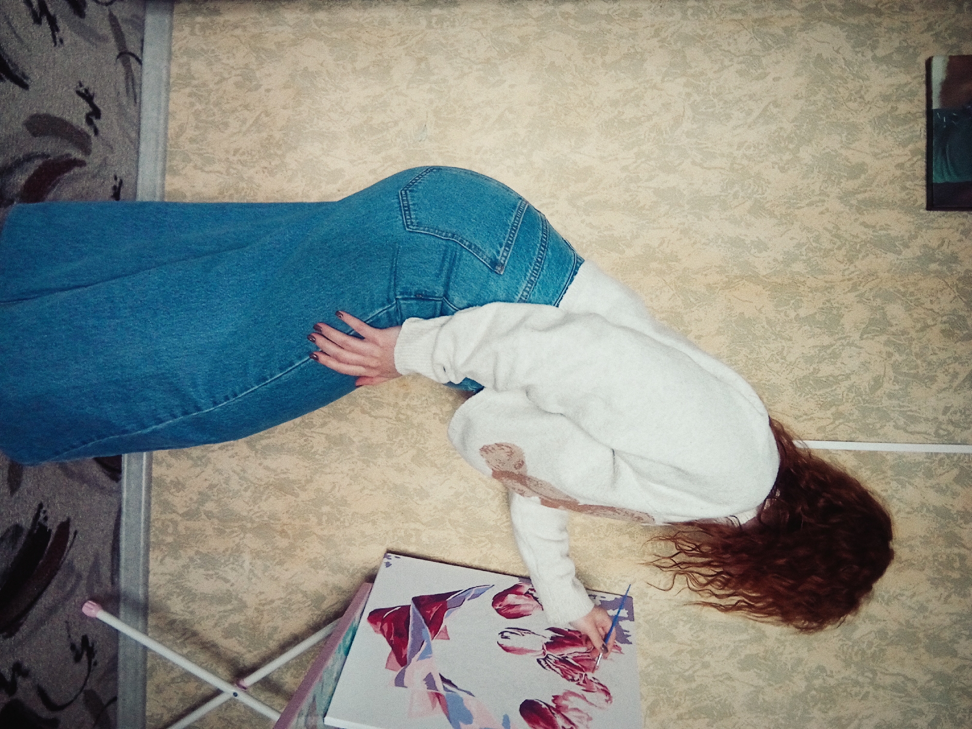 This overhead photograph captures a person engrossed in painting within an indoor setting in Yurgamysh, Russia. The subject, seen from the waist up and turned away from the camera, is a person with long, wavy, reddish-brown hair cascading towards the lower right of the frame. They are dressed in blue jeans and a light-colored (off-white or cream) long-sleeved sweater, which features a reddish-brown mark or design on one sleeve near the elbow.
The individual is actively engaged in painting on a canvas that rests on a foldable white stand or easel. They hold a thin blue paintbrush in their right hand, carefully applying paint to an artwork depicting abstract floral shapes in shades of red, pink, and purple. Their left hand gently supports the canvas or rests beside it.
The immediate surroundings consist of two distinct patterned surfaces. The dominant surface is a light beige or cream-colored floor or wall, featuring a subtle, organic, textured pattern. To the left, a darker, greyish-brown patterned rug or floor section is visible, separated by a thin vertical divider. A framed picture or artwork with dark and teal colors is partially visible on the wall in the upper right corner. The image provides a close-up, top-down perspective, highlighting the artist's focus on their creative work. There is no visible text in the image.
