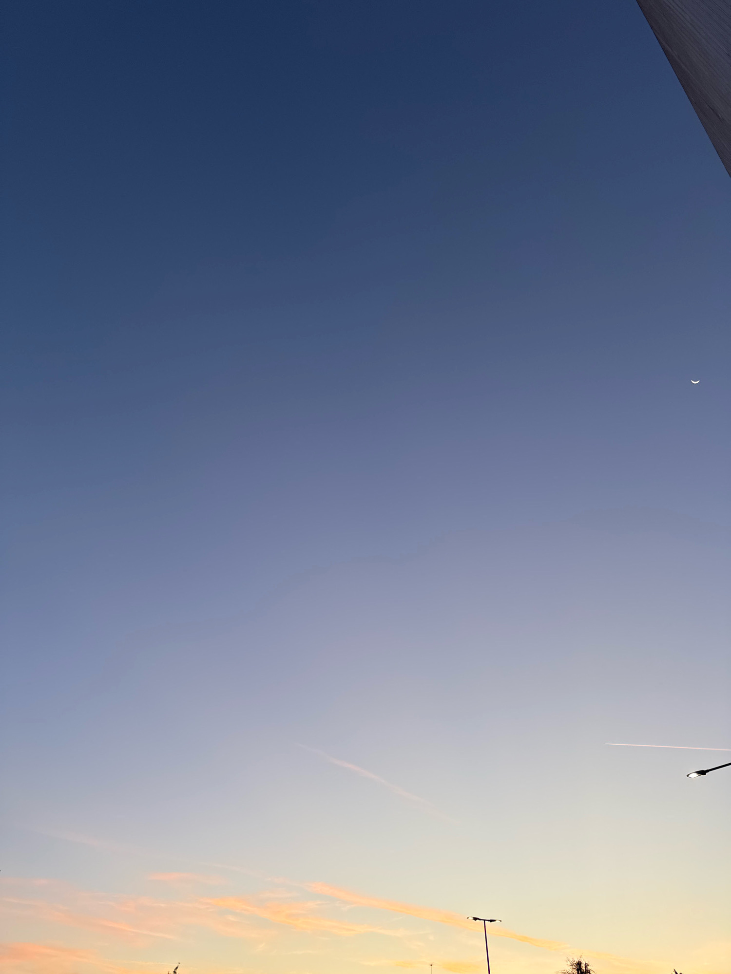 The image shows a sky at dusk with a gradient of colors from deep blue at the top to pale yellow and pink near the horizon. A thin crescent moon is visible in the upper right portion of the sky, and a faint contrail from an airplane stretches across the middle right. In the lower portion of the image, there are silhouettes of two streetlights and some bare tree branches against the colorful sky. The top right corner of the image features a dark, angled structure that appears to be a building or part of a larger construction. The scene suggests an outdoor urban environment at the end of the day. The location provided is Mejaniga, Italy.