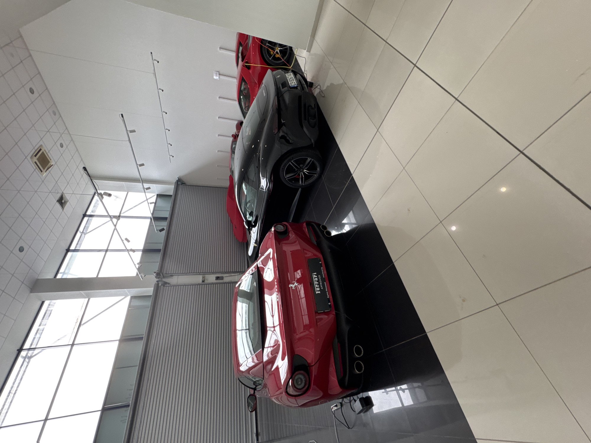 The image shows the interior of a Ferrari dealership, likely located in the city of Sofia, Bulgaria, based on the license plate of one of the cars. Three Ferrari cars are visible, lined up in the showroom. The first car, closest to the viewer, is red. The second car is black. A glimpse of a third red Ferrari car is seen behind the black one. The showroom features large windows, a tiled floor with black and white sections, and a white ceiling with track lighting. No people are visible in the frame. The environment appears modern and well-lit.
