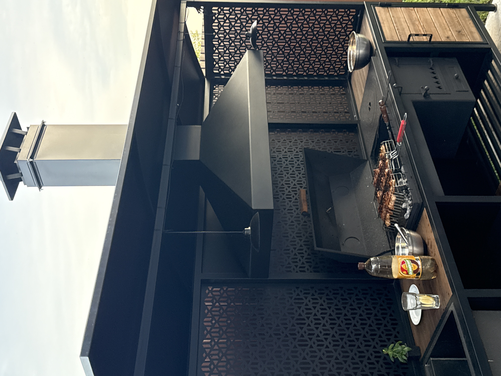 This image captures an outdoor barbecue setup in what appears to be a backyard or patio area in Starokorsunskaya, Russia. The main subject is a large, modern black metal barbecue grill structure. It features a prominent black range hood, a grilling area with a rack of skewered food, and various shelves and compartments. To the left, a tall, silver metal chimney or ventilation pipe extends upwards. On a wooden shelf to the right, there are several items: a metal bowl, a large plastic bottle of oil with a yellow and red label, a small shot glass on a saucer, and a small potted plant. Above the grill area, a decorative metal screen with a geometric pattern adds visual interest. The overall scene suggests a preparation for or the active process of grilling, indicated by the food on the skewers. The lighting suggests it is daytime, possibly late afternoon, with a bright, overcast sky visible in the background. The setting is a residential outdoor space, with wooden elements and metal construction of the barbecue unit being the most prominent features. There is no visible text other than what is on the oil bottle label, which is not clearly legible.