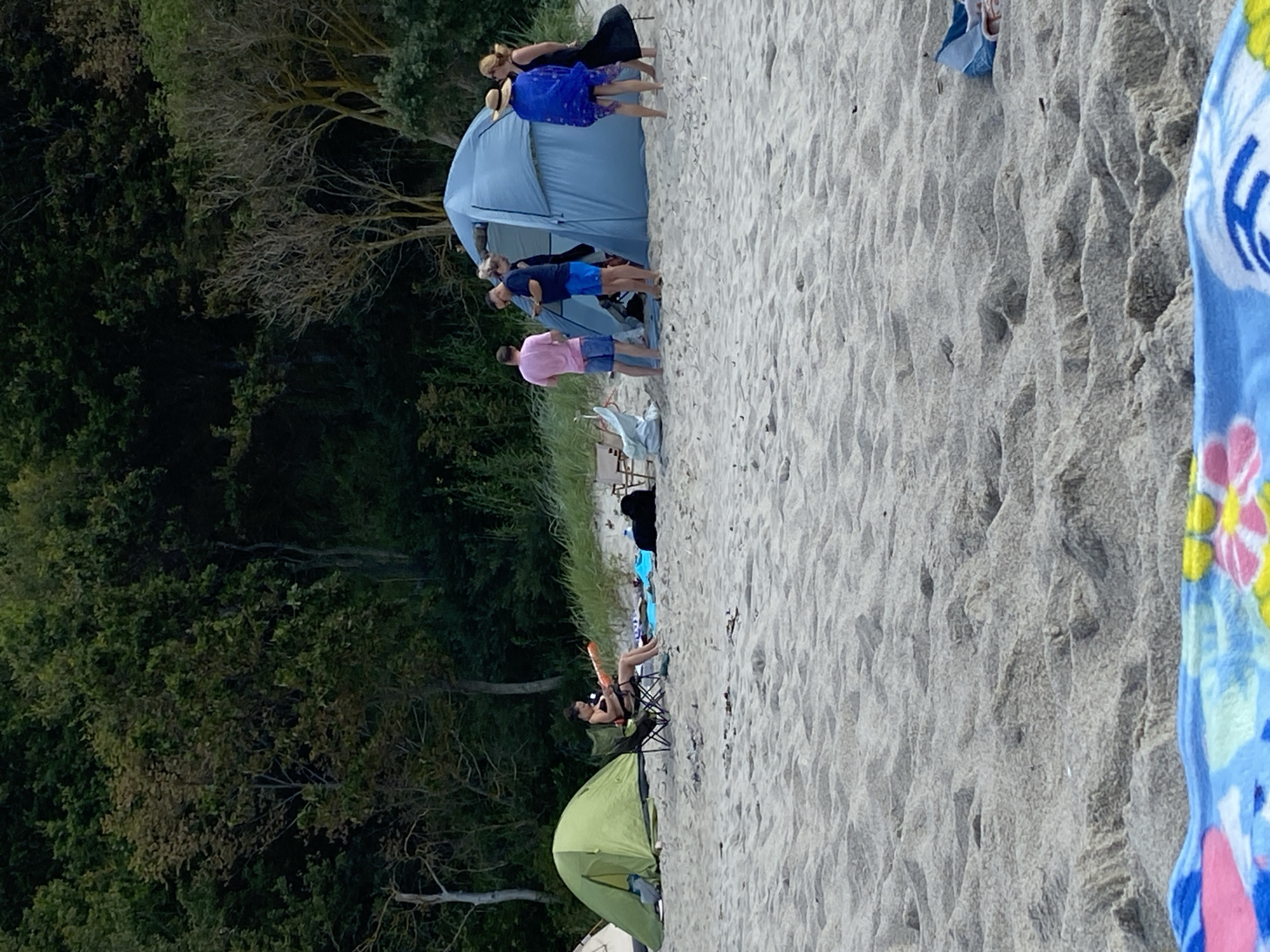 A group of people are gathered on a sandy beach with a forest in the background. In the foreground, a colorful beach towel is laid out on the sand. Further back, a woman in a black bikini sits in a camping chair next to a green tent, holding a bright orange object. To the right, a large blue tent is set up, with several people standing around it, some appearing to be talking. The environment is a beach with sand dunes and vegetation, suggesting a natural or semi-developed coastal area. The image captures a casual outdoor gathering, likely for camping or a day at the beach.