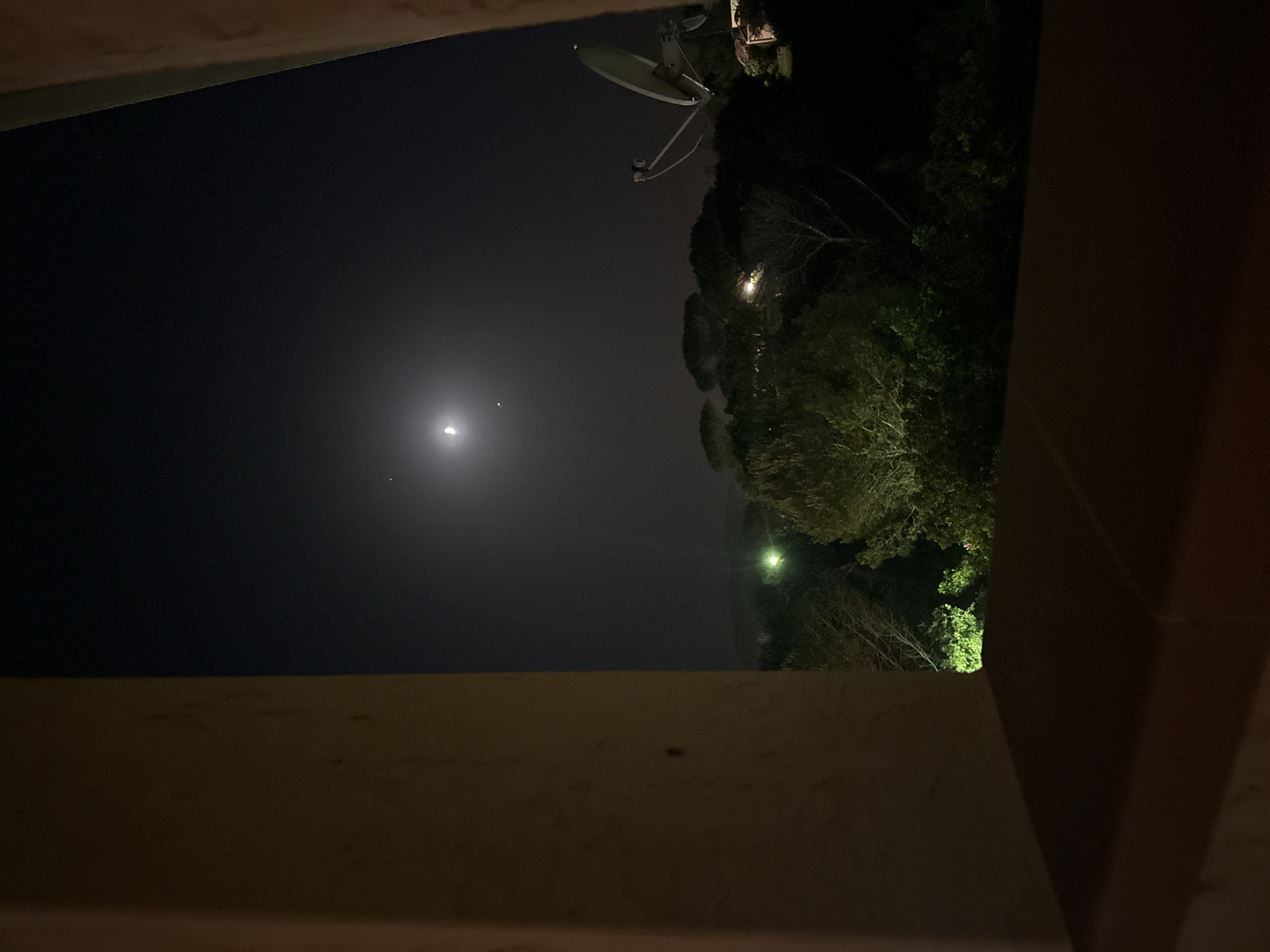 The image displays a night scene, possibly viewed from a window, looking out over a landscape. In the sky, a crescent moon is visible, accompanied by two smaller celestial bodies. Below the sky, a dark silhouette of trees and foliage dominates the middle ground. Scattered among the trees are faint, artificial lights, one of which appears bright green. On the right side of the frame, a satellite dish stands out against the dark sky and trees. The foreground is partially obscured by a warm-toned, textured surface, likely a windowsill or ledge, and a portion of a wall or pillar on the left. The overall impression is of a rural or semi-rural environment at night, potentially in or near Biddeputzi/Villaputzu, Italy, given the context. No people or specific actions are depicted.