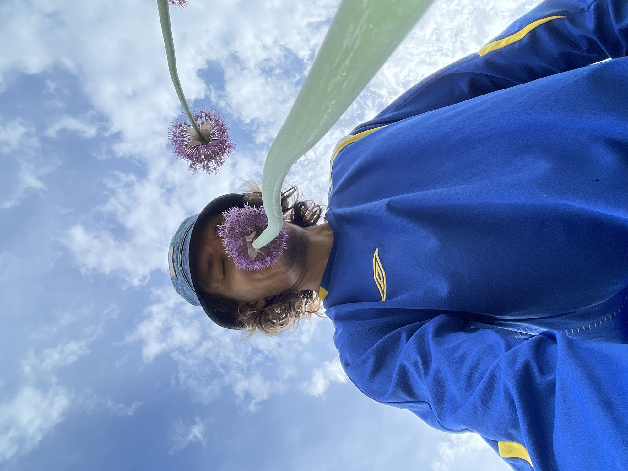 A low-angle shot shows a person with curly hair, wearing a blue and yellow athletic jacket and a hat, looking down at the camera from under a sky with scattered clouds. Two allium flowers on green stems are prominently featured; one is positioned near the person's face and the other above. The person is located in the city of Перевозное, Russia.