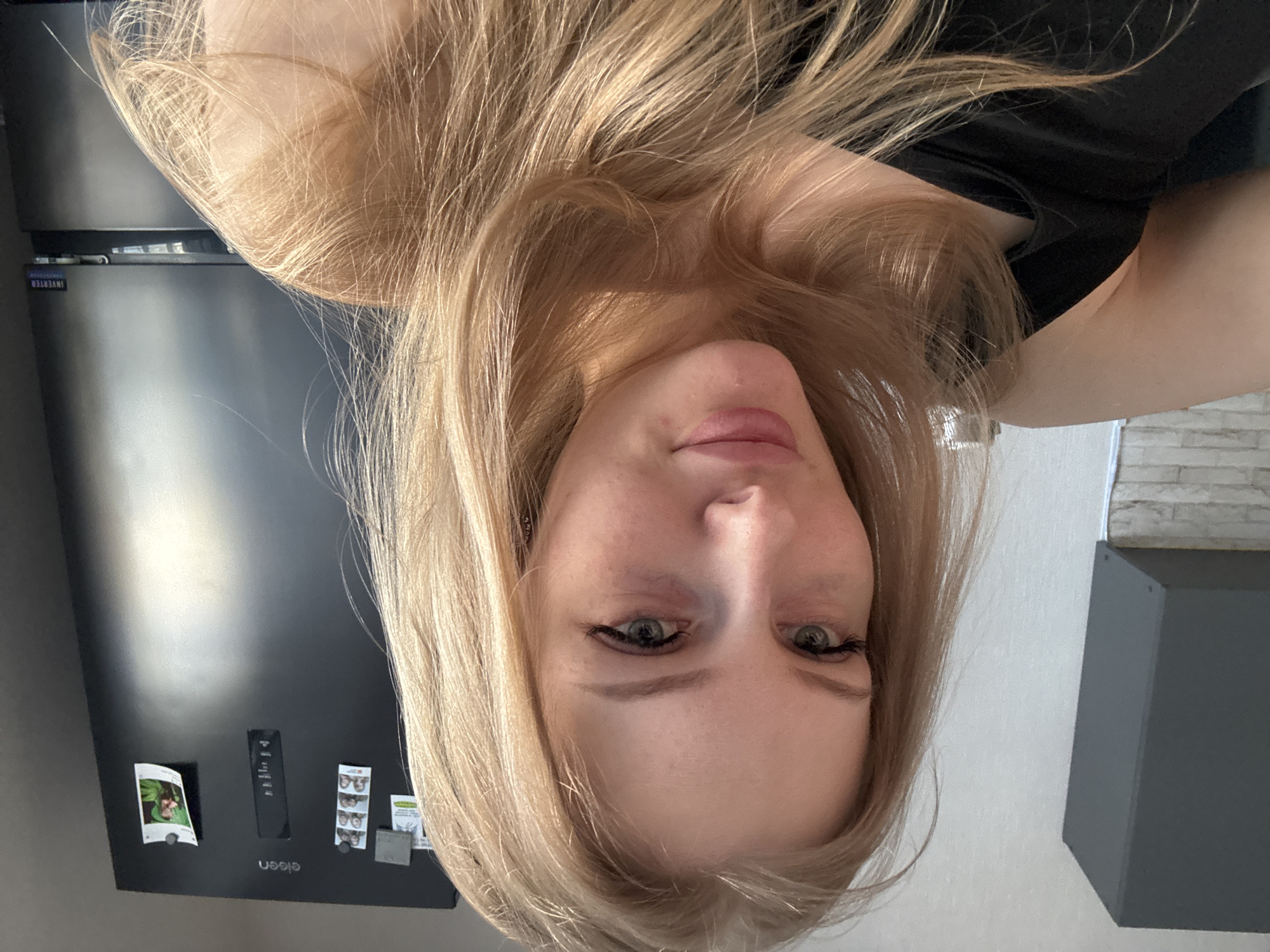 A woman with blonde hair is pictured indoors. She is wearing a black shirt and appears to be in a kitchen, as a black refrigerator is visible behind her. The refrigerator has several magnets and notes attached to it. A gray box is also in the background near a white brick wall. The location is assumed to be Perm, Russia.