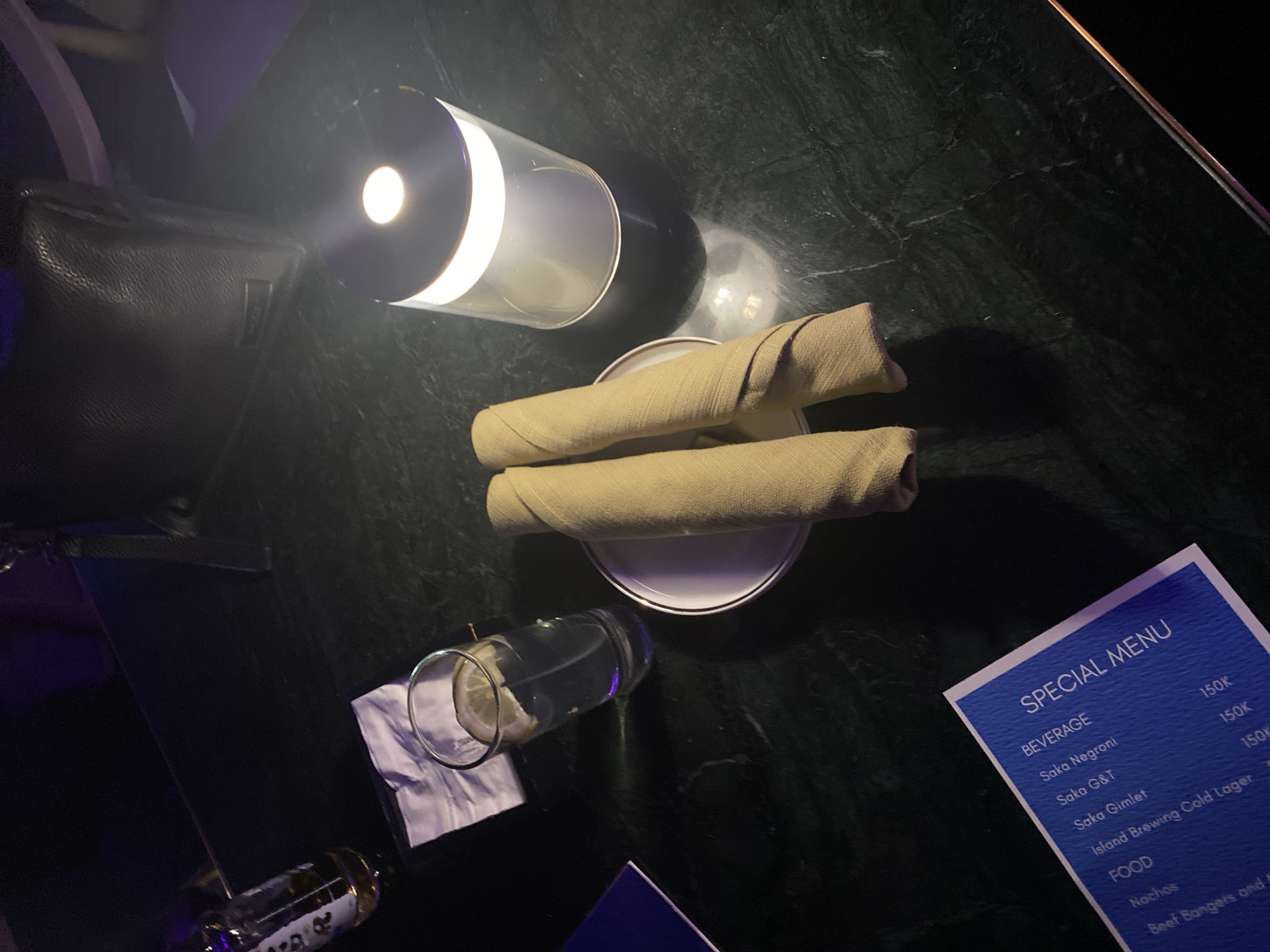 The image captures a dimly lit, upscale table setting, likely in Central Jakarta, Indonesia. The focal point is a dark, polished table surface, possibly marble, with a slight sheen. On the table, a clear glass filled with water and a lemon slice sits beside a neatly folded white napkin. Adjacent to the glass is a white plate with two rolled-up, light brown napkins resting on it. A modern, cylindrical LED lamp with a bright white light is positioned to the right, casting a warm glow and a circular reflection on the table. In the background, a dark, textured handbag is visible, and further back, hints of purple and dark lighting suggest a bar or lounge environment. A portion of a blue menu is visible in the lower left corner, displaying text that includes "SPECIAL MENU," "BEVERAGE," "Saka Negroni," "Saka G&T," "Saka Gimlet," and under "FOOD," "Nachos" and "Beef Banger and M". The overall ambiance suggests a sophisticated evening out.