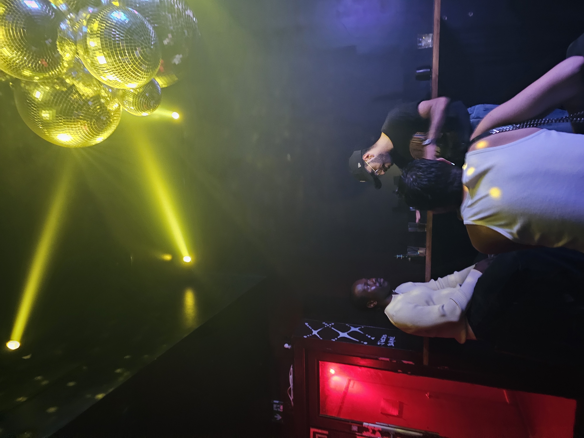 The image shows the interior of a club or bar, likely in Paris, France, based on the upscale, modern aesthetic and potential clientele. In the upper left, there is a cluster of disco balls illuminated by yellow spotlights, creating a dynamic light effect across the scene.
Three people are visible, suggesting social interaction in the bar/club setting. A person in a black shirt and a baseball cap appears to be near or behind the bar. A person wearing a white tank top is also behind the bar. A third person is wearing a light-colored sweater and dark pants, standing further back in the image.
In the background is a red lighted door. There are shelves above the door.