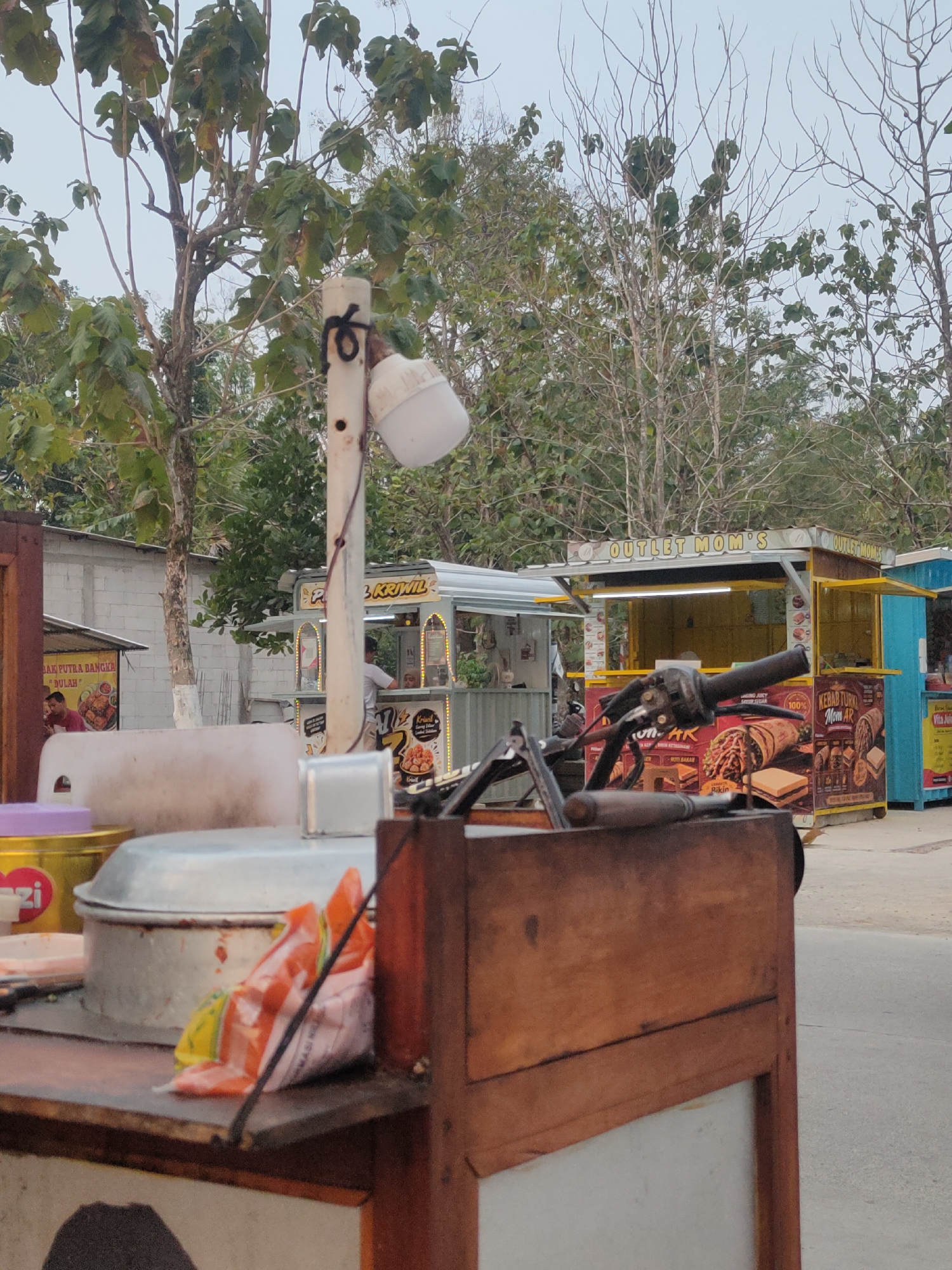 The image depicts an outdoor scene at what appears to be a street food market or roadside vendor area in Karanglangu, Indonesia. In the foreground, a portion of a wooden food cart is visible, with various food items and containers on its surface, including plastic bags of snacks and a metal pot. Behind the cart, a motorcycle handlebar is prominent. Further in the background, several food stalls are set up. One stall has signage that reads "OUTLET MOM'S" in large letters and displays pictures of food items. Another stall, to the left, has a sign with Indonesian text and images of fried chicken. The stalls are brightly lit, and there are some people visible inside or near them, seemingly customers or vendors. The environment suggests a commercial area with trees and a clear sky.