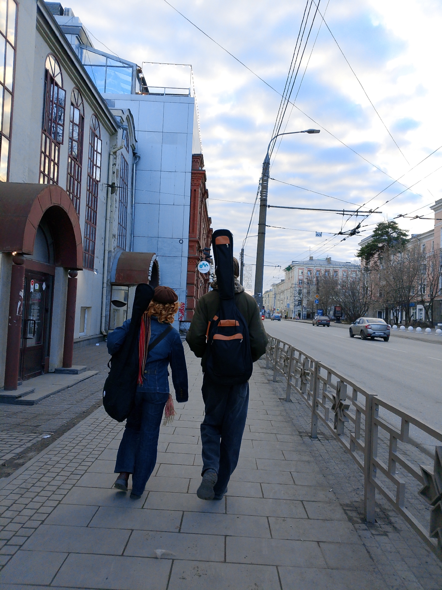 Two people with guitar cases are walking on a sidewalk. The person on the left is wearing a denim jacket and the person on the right is wearing a dark jacket and a backpack. They are walking away from the viewer. A gray car is driving on the street next to the sidewalk. The location is Izhevsk, Russia.