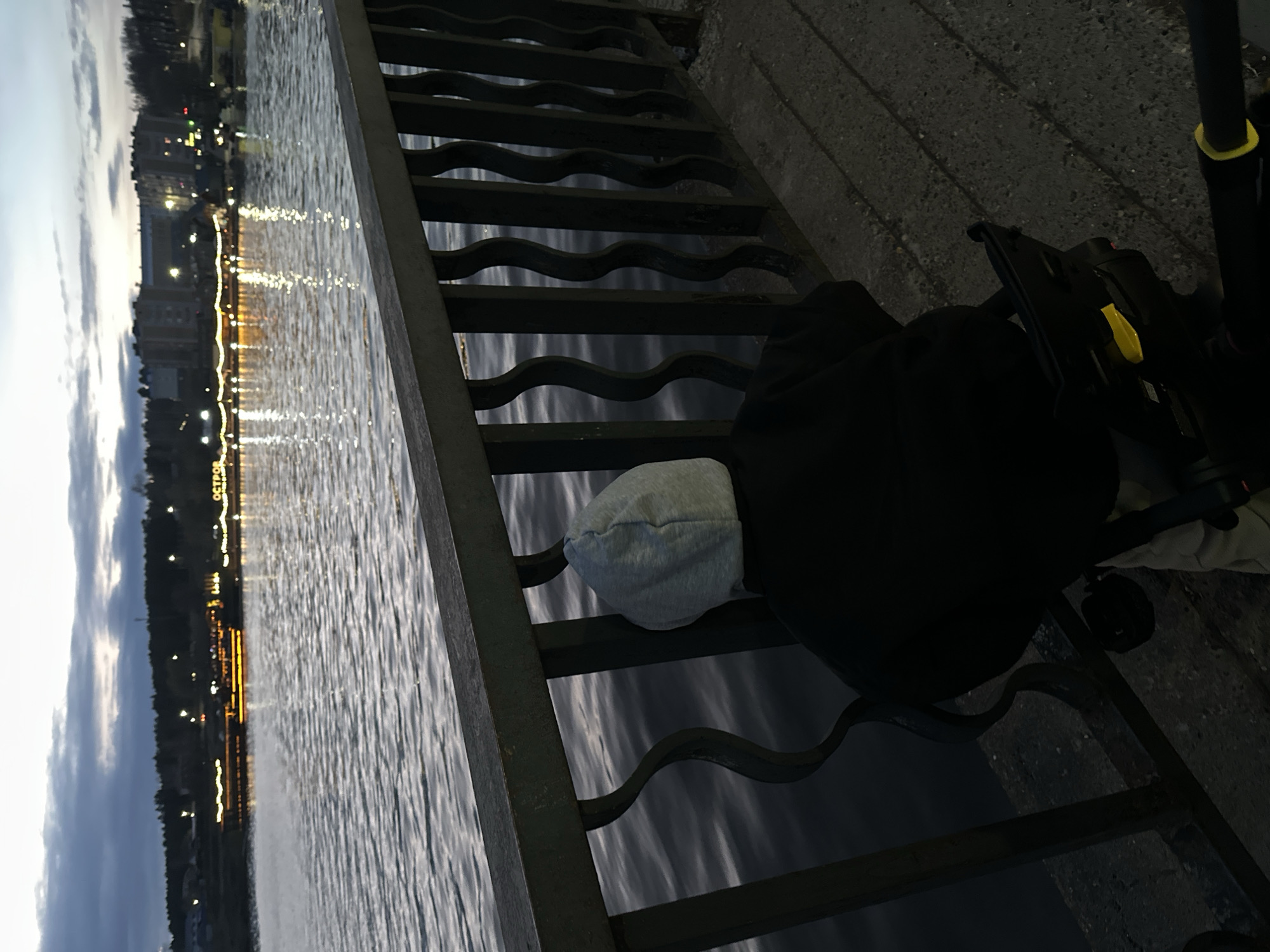 The image, captured at dusk or twilight, depicts a serene scene overlooking a body of water in Votkinsk, Russia. A person, viewed from behind, stands beside a dark metal railing with decorative wavy bars, appearing to gaze out across the water. They are dressed in a black hooded top and a light grey beanie or hood. Adjacent to the person on the right, part of a black and yellow stroller or similar wheeled object is visible.
The water, likely a river or lake, is calm with numerous bright reflections from city lights on the opposite bank. These luminous streaks create a striking contrast against the dark, rippling surface of the water. The sky above is dim, showing subtle cloud formations.
On the distant bank, a line of buildings and trees is illuminated, with their lights dancing on the water. Notably, some Cyrillic text is visible on the far bank, spelling out "ОСТРОВ" (meaning "island"), likely a sign for a building or landmark. The ground where the person stands appears to be a textured concrete surface. The overall atmosphere of the scene is peaceful and contemplative, capturing a quiet moment by the water as day transitions into night.