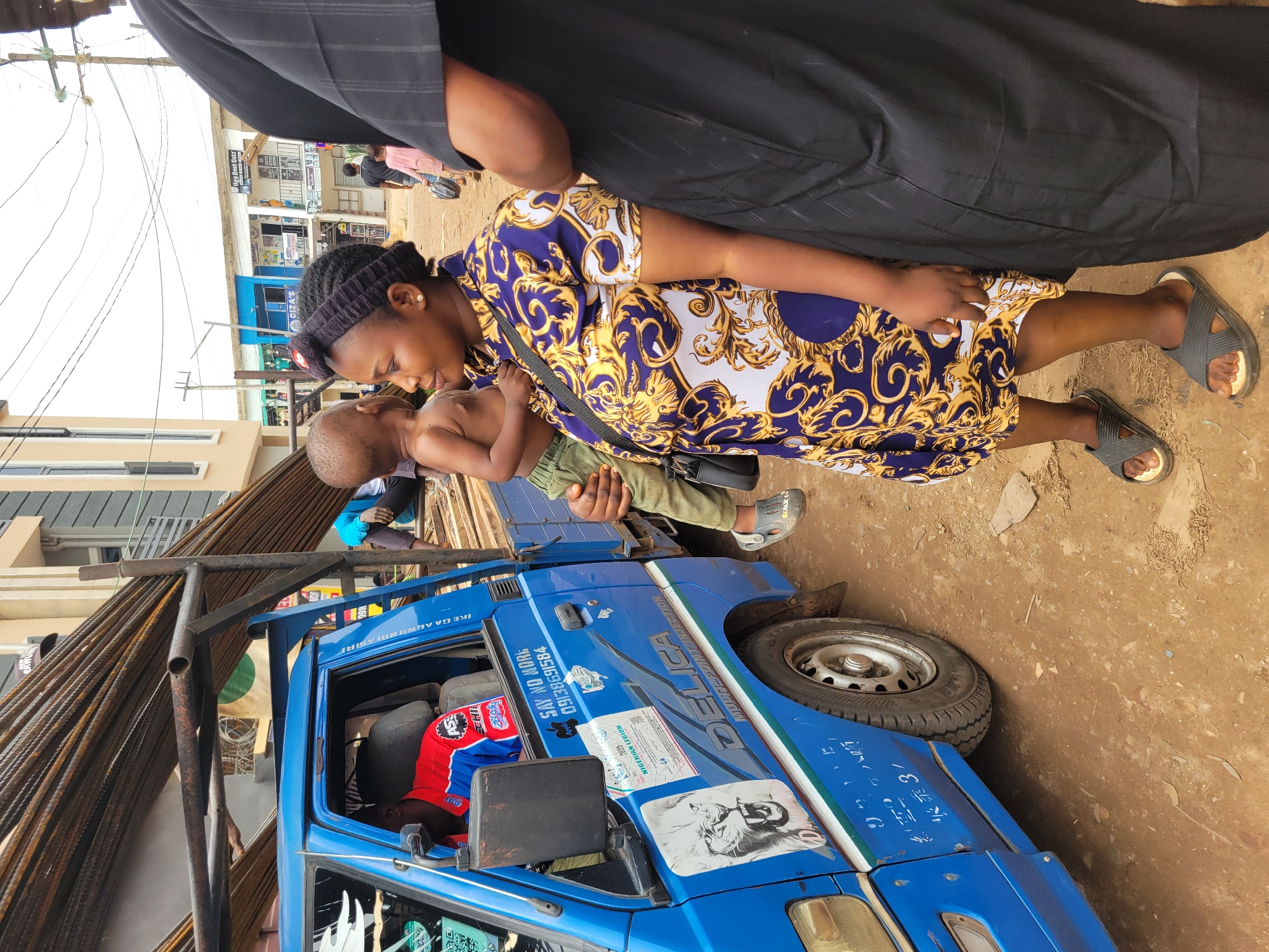 In this medium shot, a woman in a vibrant blue and gold patterned dress holds a young child. She is looking down at the child, their faces close together, suggesting a tender moment. The woman wears a black headband and grey sandals. The child, partially visible, appears to be wearing green trousers. They are positioned in front of a bright blue truck loaded with long metal rods.
The truck has various stickers and text on its side, including "DELICA MULTI POINT INJECTION," "SAY NO MORE 09138691584," and a Nigerian Legion sticker with the year "2025." The word "Apple" is also visible on a sticker. The scene is outdoors, with a dirt or gravel ground. In the background, buildings and power lines are visible, indicating an urban or semi-urban setting. The overall impression is a candid snapshot of daily life in what appears to be a bustling marketplace or street. The lighting suggests it could be daytime.