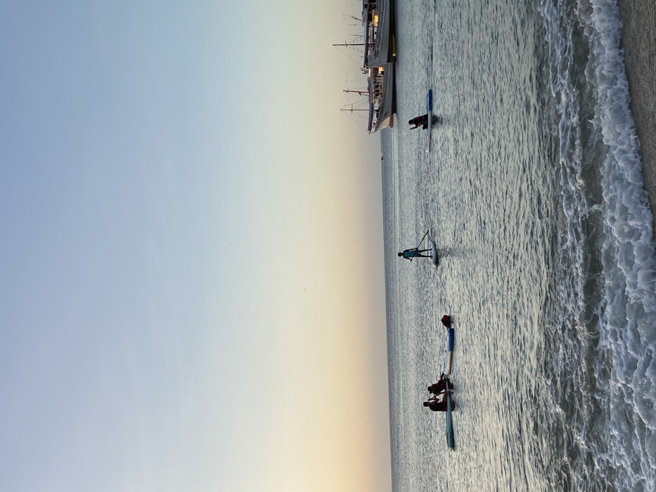 A tranquil beach scene unfolds at dusk. Gentle waves lap against the sandy shore. In the calm water, individuals are paddleboarding, their silhouettes visible against the fading light. A large boat rests further out in the water, possibly anchored, adding to the serene coastal ambiance. The sky transitions from blue to a soft yellow hue.