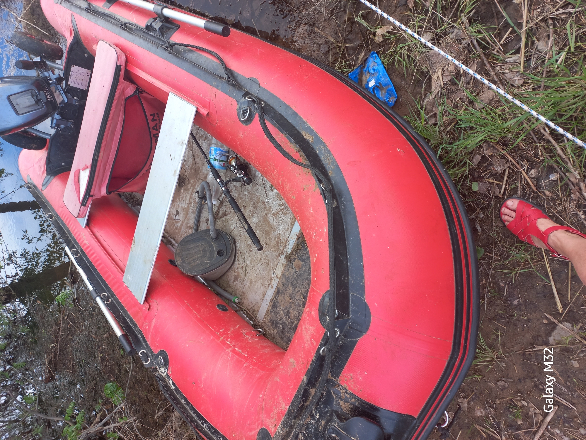A red inflatable boat rests partially in the water and partially on the shore. The boat is equipped with a small outboard motor and oars. Various pieces of equipment, including fishing rods, are inside the boat. A person wearing a red sandal is visible near the edge of the boat. The surrounding environment consists of grass, mud, and trees. The photo was taken on the banks of the Akhtuba River in the city of Volzhsky, Russia.