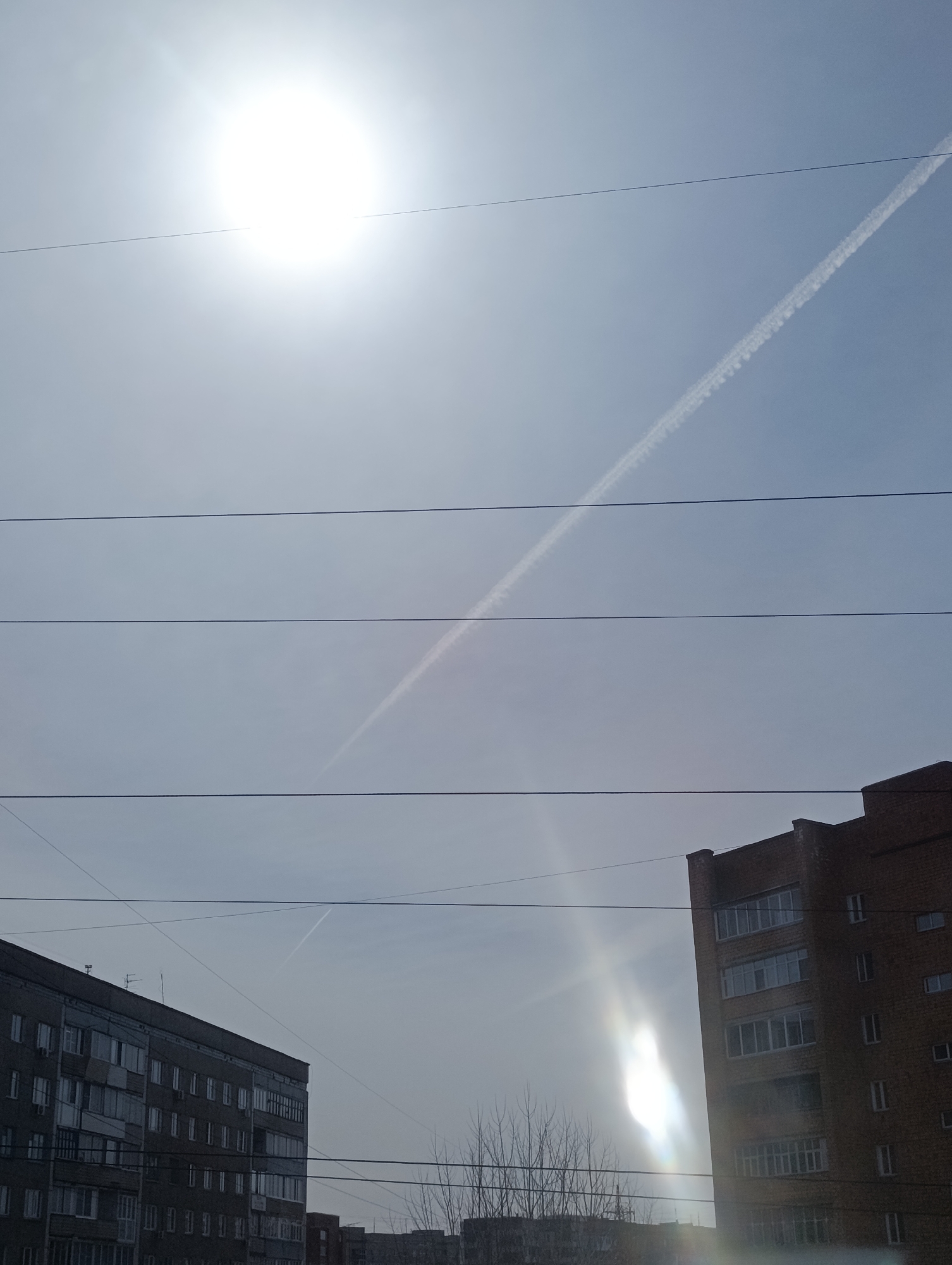 The image shows a daytime scene in Krasnoyarsk, Russia. The sky is overcast with a bright sun high in the frame. A contrail stretches across the sky, intersecting multiple power lines. Two multi-story apartment buildings flank the scene. The building on the left is gray, while the building on the right is brick. A glare from the sun is visible between the building on the right and bare trees.