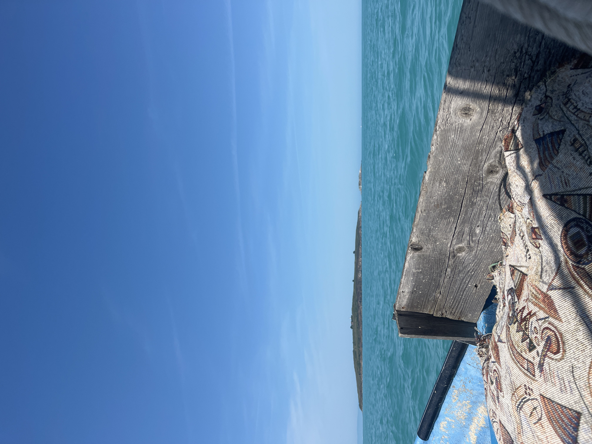 The scene depicts a view from a watercraft in a large body of water, possibly an ocean or sea, at an unspecified location in Unknown, Unknown. The upper half of the image features a clear, vivid blue sky with minimal, high-altitude wispy clouds. Below the horizon line, the water is a vibrant turquoise, exhibiting small surface ripples and waves.
A low-lying, elongated island, densely covered with green vegetation, occupies the mid-distance on the left side of the frame. Faintly visible on its peak are one or two small, light-colored structures. A much smaller, possibly rocky islet is discernible further to the right on the horizon line.
In the immediate foreground, the edge of a boat is visible. To the left, a portion of a blue-painted boat hull, showing significant wear, scratches, and paint flaking, is present. A black cylindrical object, possibly a fender or part of the boat's structure, is mounted horizontally along the interior of the blue hull. Dominating the lower right is a weathered, grey wooden plank or beam, serving as a railing or structural component. The wood is coarse-grained, with visible knots and splits, indicating age and exposure. A strong, dark, diagonal shadow falls across this wooden plank and the patterned, woven fabric directly below it. The fabric, which appears to be a cushion or covering, displays an intricate, abstract design in earthy tones (brown, beige, reddish-orange), featuring shapes that resemble stylized fish, birds, or nautical symbols. The fabric appears slightly rumpled and worn. No people or animals are visible in the scene.