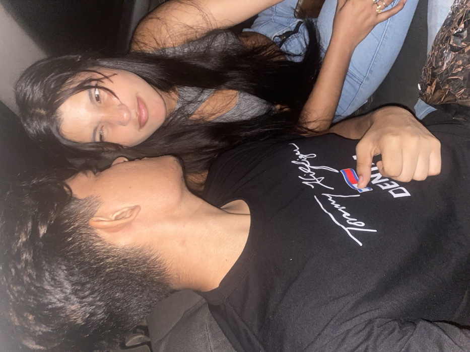 A young woman with long dark hair, wearing a gray tank top and light blue jeans, is lying on top of a young man. She is gazing at him with a slightly serene expression. The man, wearing a black Tommy Hilfiger shirt, has his head turned slightly to the side. They appear to be close, possibly a couple. The lighting is subdued, casting a soft glow on their faces.