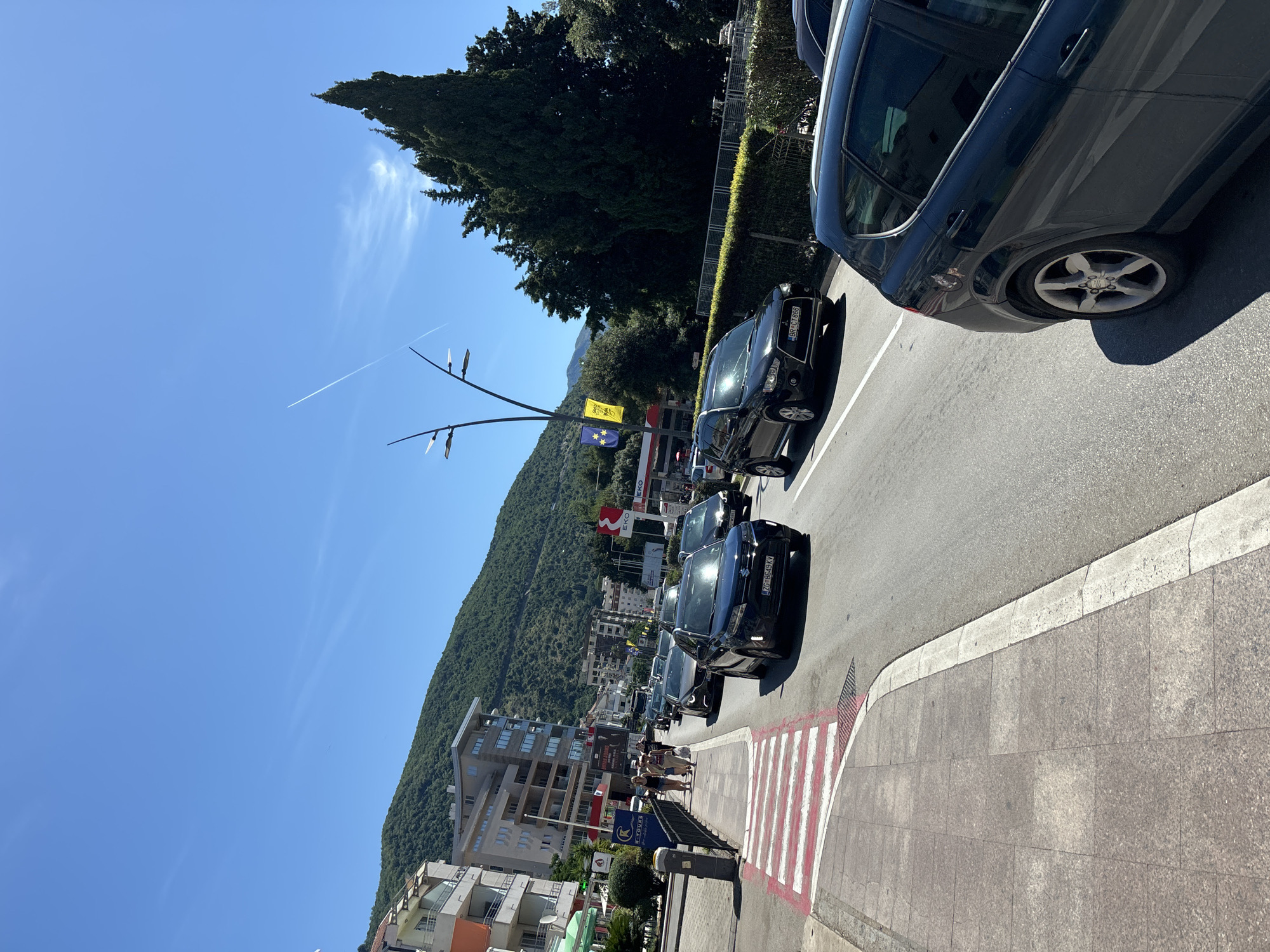 The image captures a street scene in Budva, Montenegro, on a clear, sunny day. A group of pedestrians is walking on a sidewalk adjacent to a busy street filled with cars, indicating moderate traffic. The architecture features several multi-story buildings, characteristic of a coastal town. In the background, a lush, green mountain slopes upwards, providing a natural backdrop to the urban environment. The sky is a vibrant blue with a faint contrail from an aircraft. The street is paved, with a pedestrian crossing marked with red and white stripes leading to the sidewalk. Streetlights and vegetation, including a large cypress tree, are also visible, adding to the overall ambiance of the location. The presence of what appears to be a gas station with EKO branding suggests commercial activity in the area.