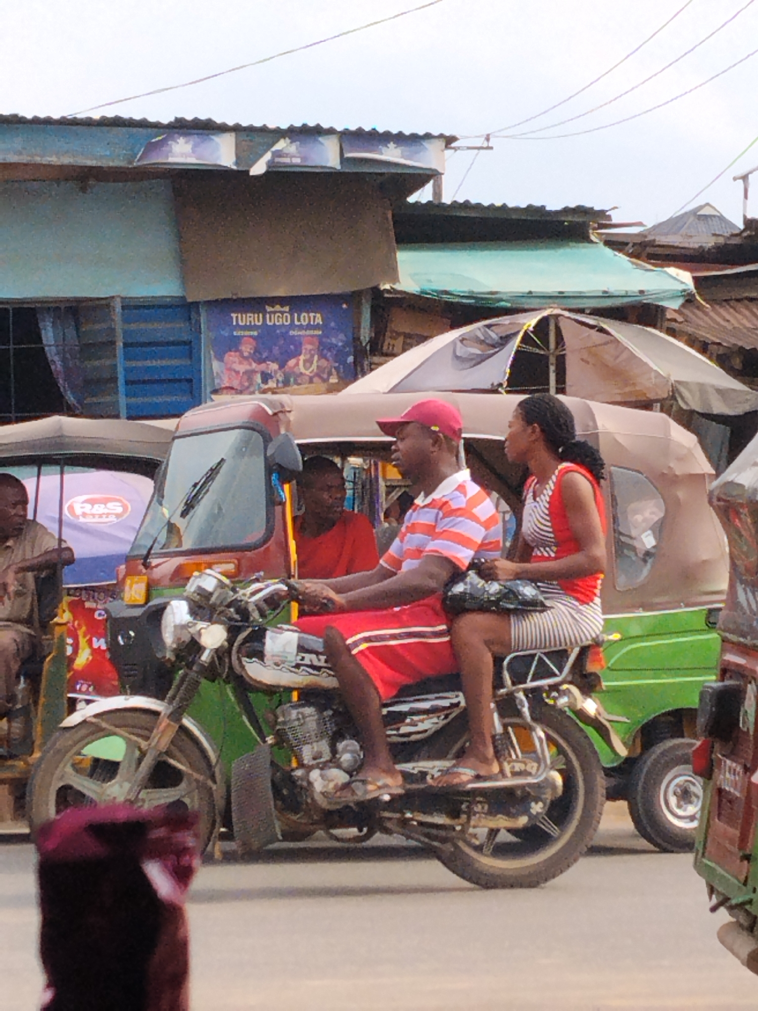The image captures a bustling street scene in Aba, Nigeria, featuring various modes of local transportation and roadside structures under an overcast sky.
In the foreground, a man in a red baseball cap, striped t-shirt, and red shorts rides a motorcycle. He wears flip-flops. Behind him, a woman with braided hair, dressed in a red and white striped dress, sits as a passenger, holding a black bag. Both riders are positioned centrally, moving or stopped amidst traffic.
To their left and behind, a red and brown tricycle, commonly known as a keke napep, is visible. A man in a red t-shirt is seated in its passenger compartment. Further left, parts of another tricycle, blue and white, are seen with the logo "R&S LOTTO" prominently displayed on its rear canopy. A man in a light-colored shirt is seated beside it. Behind the motorcycle and extending to the right, a green tricycle is visible, with its rear wheel and part of its cabin in view. An open, light-colored market umbrella is partially visible above the green tricycle, suggesting market activity.
The background consists of various buildings with corrugated iron roofs, indicative of urban commercial or residential areas. One building, with blue siding, has a billboard-like poster on its facade. This poster features the text "TURU UGO LOTA" at the top, along with smaller text "EXTREME" and "D&M BRAND." Below the text, two stylized figures are depicted, possibly promoting a product or event. Power lines crisscross the top of the frame against the bright, diffused sky.
The scene depicts daily life and transportation on a street, likely during the daytime, with no strong shadows indicating a bright sun. People appear to be engaged in routine activities, moving through the urban environment of Aba.