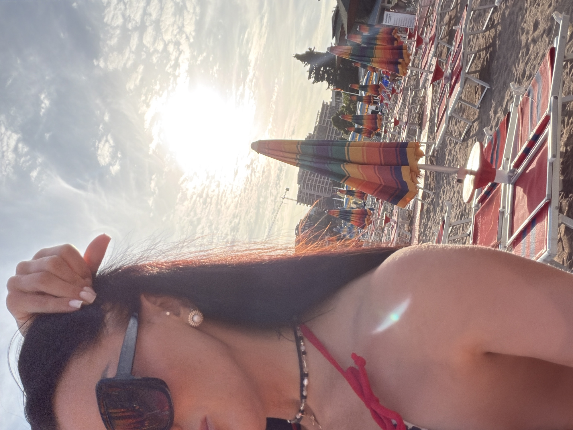 The image displays a beach scene in Boreti, Montenegro, characterized by strong backlighting from an overhead sun against a partly cloudy sky. The foreground features the left profile of a female subject, cropped from the nose/mouth to the upper arm. She wears large, dark-framed sunglasses, a pearl-and-gold stud earring with an additional small piercing above it, and a multi-beaded black choker necklace. Her dark hair is pulled back, with her right hand visible lifting a section of hair near her temple, revealing light-colored fingernails. She is wearing a red bikini top. The sandy beach is populated with numerous rows of red and white lounge chairs and multi-colored striped beach umbrellas. In the background, several multi-story buildings are visible, one with a construction crane, alongside a large coniferous tree.