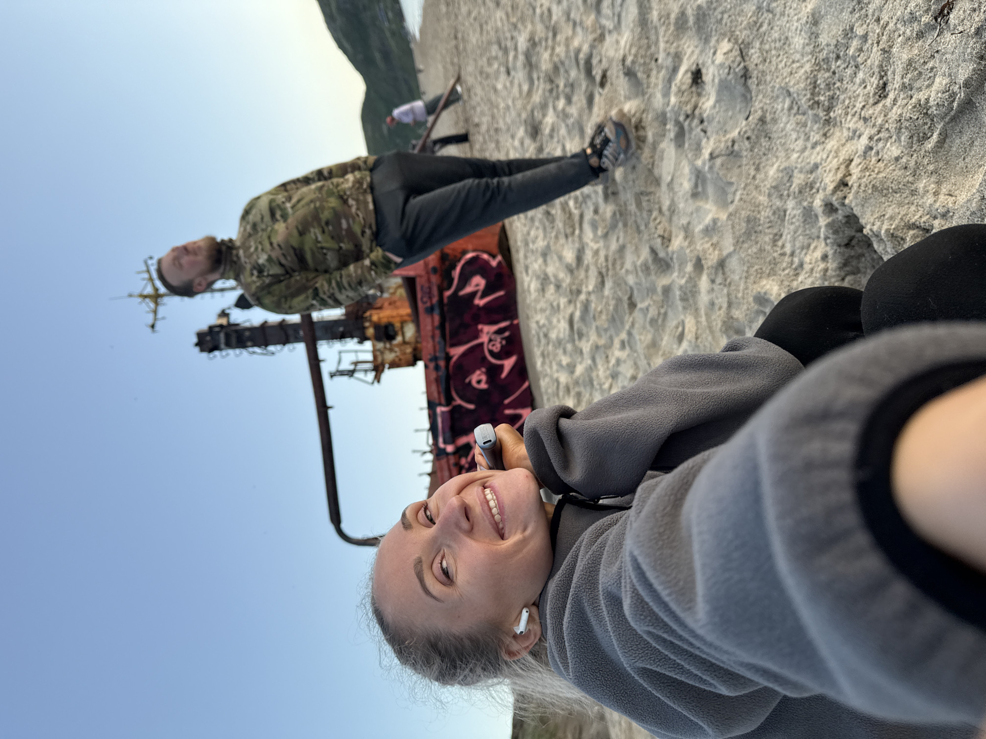 The image depicts a beach scene in Teriberka, Russia, featuring a rusted shipwreck as a prominent element. In the foreground, a woman with blonde hair pulled back is smiling and looking at the camera, holding a white object to her cheek. She is wearing a gray fleece jacket and white wireless earbuds. Behind her, a man with a beard, wearing a camouflage jacket and dark pants, stands with his hands in his pockets, looking upwards. In the background, more people are visible walking on the sandy beach, with hills and the ocean in the distance. The sky is clear and blue, suggesting daylight. The shipwreck, covered in graffiti, is partially submerged in the sand.