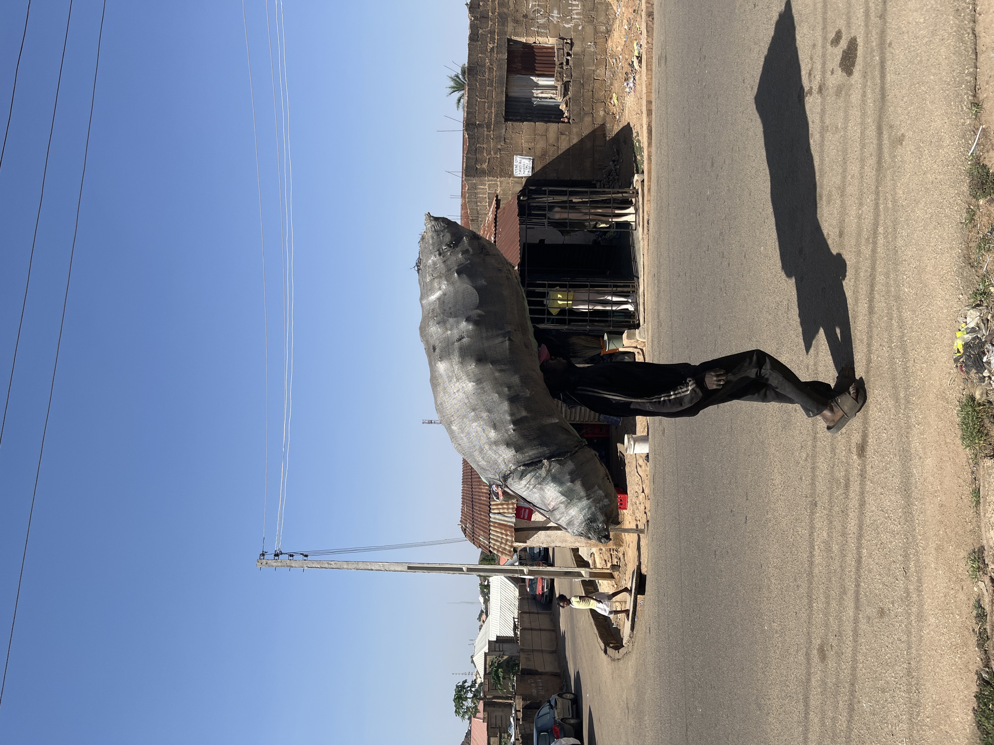 A street scene in Jos, Nigeria is depicted. A man carries a large, dark gray sack on his head, walking along the right side of an asphalt road. He wears dark pants with a white stripe and brown shoes. The road is bordered by a building with a caged storefront, and another taller building further back. Power lines run parallel to the road. The sky is a clear blue. The man's shadow stretches out onto the road.