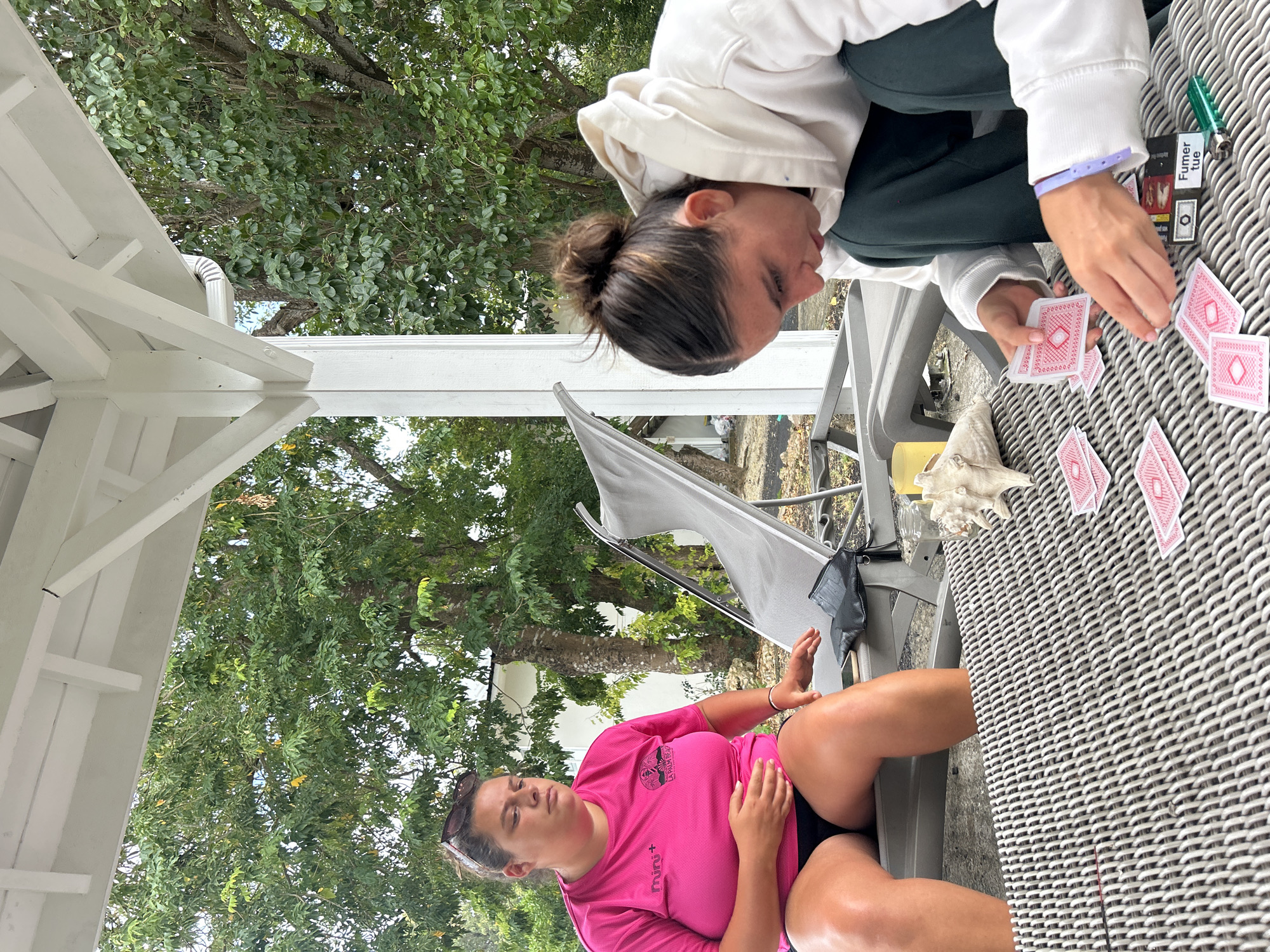 Two women are playing cards at a gray wicker table on a covered patio with white columns. One woman, wearing a pink shirt, sits on a chair while the other, wearing a white hooded sweatshirt, leans over the table, examining cards. Additional cards are scattered on the table, along with a lighter and a pack of cigarettes labeled "Fumer tue." A large tree and glimpses of a white building are visible in the background. Based on the cigarette package, this image is likely taken in France, but the exact location in Sainte-Anne cannot be determined from the visual information alone.
