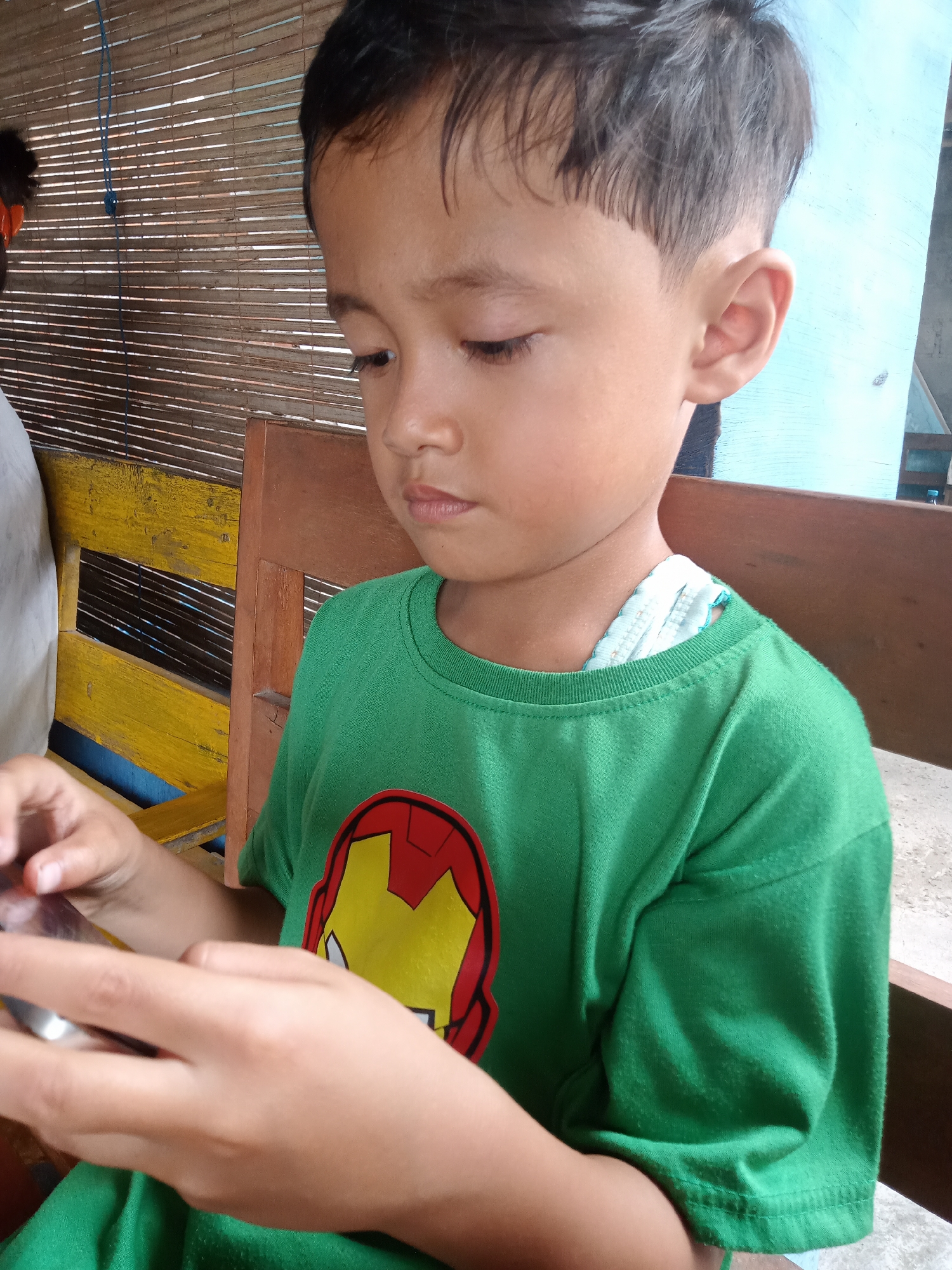 A young boy, likely of Southeast Asian descent, is depicted in a close-up, chest-up shot, looking down intently at a smartphone he holds with both hands. He has short, dark hair and fair skin. He is wearing a green short-sleeved t-shirt featuring a prominent red and yellow Iron Man mask logo on the chest. A piece of light blue and white fabric is visible underneath his t-shirt collar, suggesting another garment or cloth.
The boy is engaged in an activity on the smartphone, his eyes focused downwards, indicating concentration. His expression is calm and absorbed. The setting appears to be a casual indoor or semi-outdoor area in Bungur, Indonesia. The background features a textured wall made of horizontal wooden or bamboo slats. To the left, part of a yellow-painted wooden bench or chair back is visible, along with a blue string hanging down. To the right, a darker brown wooden arm or backrest of a chair can be seen, behind which is a light blue wall or surface and a concrete floor. The lighting suggests it is daytime. No other distinct text is visible apart from the Iron Man logo.