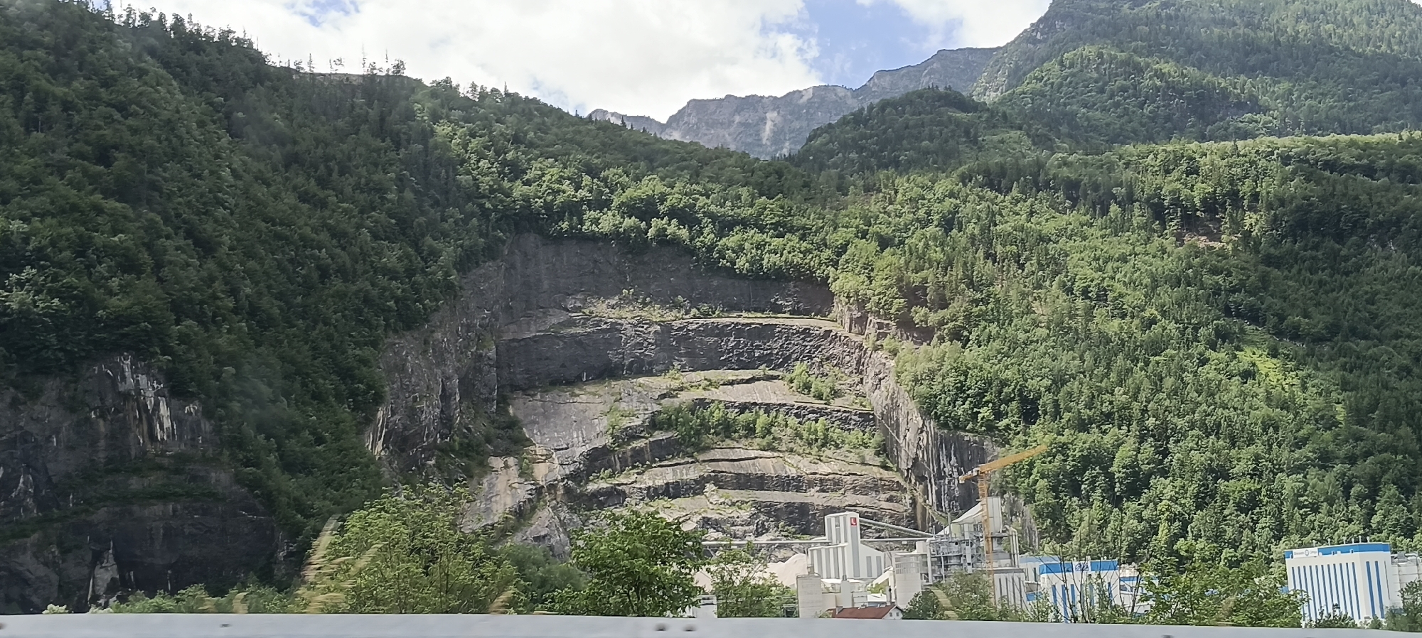 The image shows a quarry in the mountainside, located in Torren, Austria. The quarry is characterized by a series of horizontal rock layers that have been excavated, creating distinct terraces. Dense green forests cover the slopes above and around the quarry, with some patches of rock visible on the steep faces. In the foreground, a modern industrial complex is situated at the base of the mountain, featuring several white and grey buildings, storage silos, conveyor belts, and a yellow crane. The sky above is partly cloudy, with patches of blue visible. A white barrier runs along the bottom of the image, suggesting a road or viewpoint.