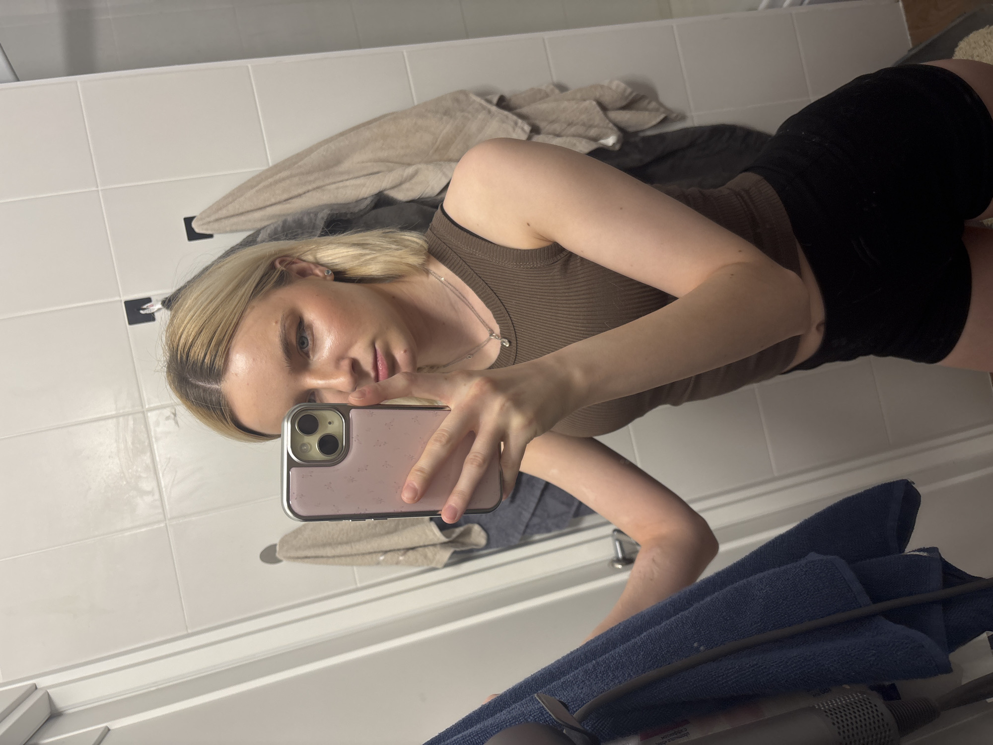 A mirror selfie captures a young woman standing in what appears to be a bathroom. The image is oriented sideways, suggesting the original photo was taken in portrait mode and then rotated.
**Subjects:** The primary subject is a young woman with blonde hair, styled back with visible darker roots, and a light complexion. She is wearing a fitted, brown ribbed tank top and black shorts. A delicate silver necklace with a small pendant and a stud earring in her left ear are visible. She is holding a smartphone with a light pink case, featuring a subtle pattern, to take the photo. Her expression is neutral as she looks directly into the phone's camera, reflected in the mirror.
**Scene and Setting:** The background reveals a white tiled wall behind the woman. To her left, draped over what seems to be a hanger or hook, are light beige and grey towels or clothing items. Below the mirror, a white ledge or edge of a fixture (possibly a bathtub or sink) is visible. A dark blue towel and part of a Dyson hair dryer are resting on this surface in the foreground.
**Activity:** The woman is actively taking a mirror selfie, holding her phone up to capture her reflection.
**Notable Details:** The lighting appears to be artificial indoor lighting. No specific time of day or weather cues are evident. The mirror reflection clearly shows the phone she is using for the selfie. There is no clearly visible or legible text in the image. The overall setting suggests a private, domestic space like a bathroom.