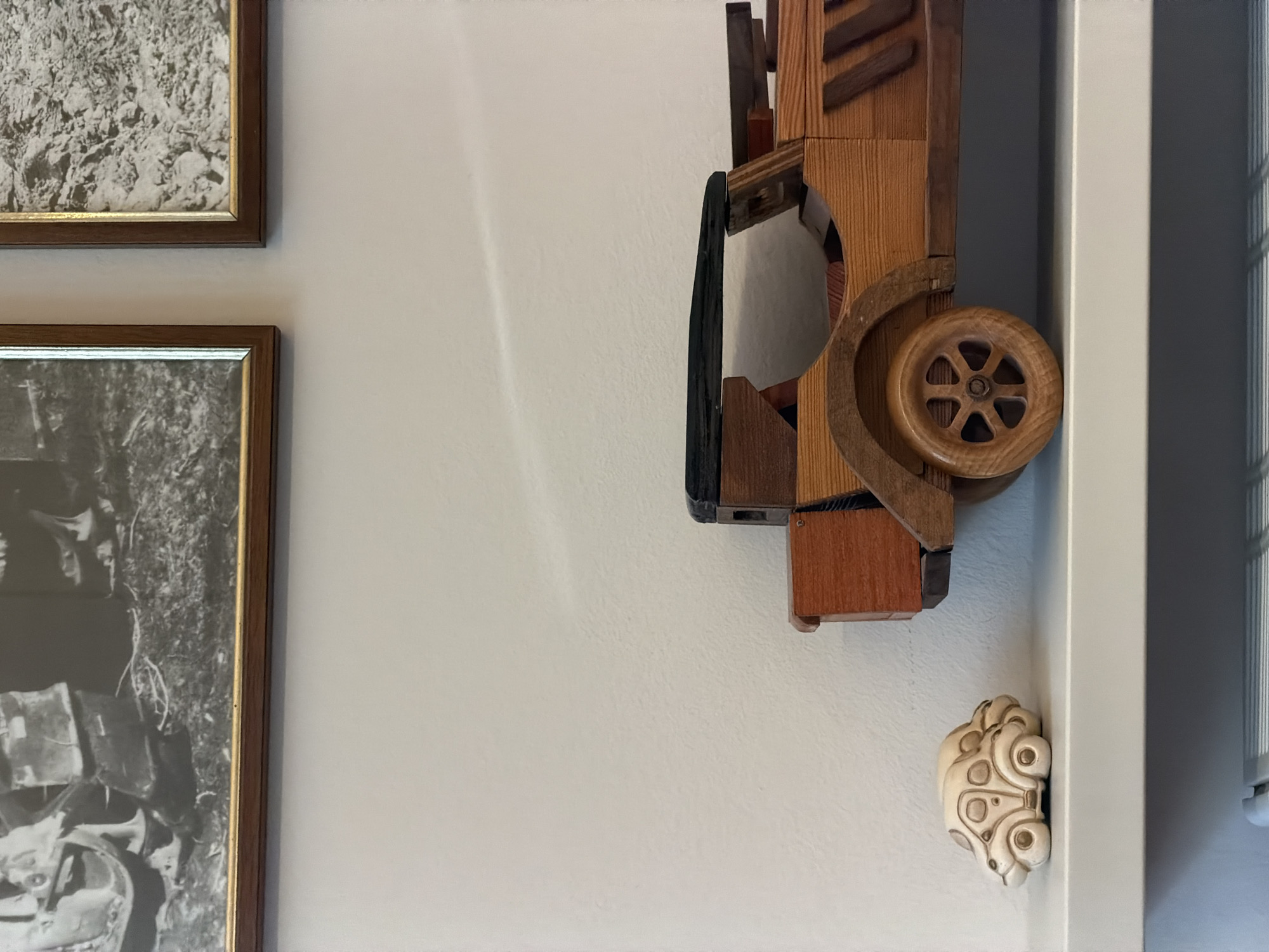 Two framed black and white photographs are mounted on a light-colored wall. Below the photographs, on a white shelf, are two toy cars. To the left is a small, light-colored ceramic car. To the right is a larger wooden toy car with a black roof and wooden wheels. The scene is likely an indoor setting, possibly a home or a display area in Arco, Italy.