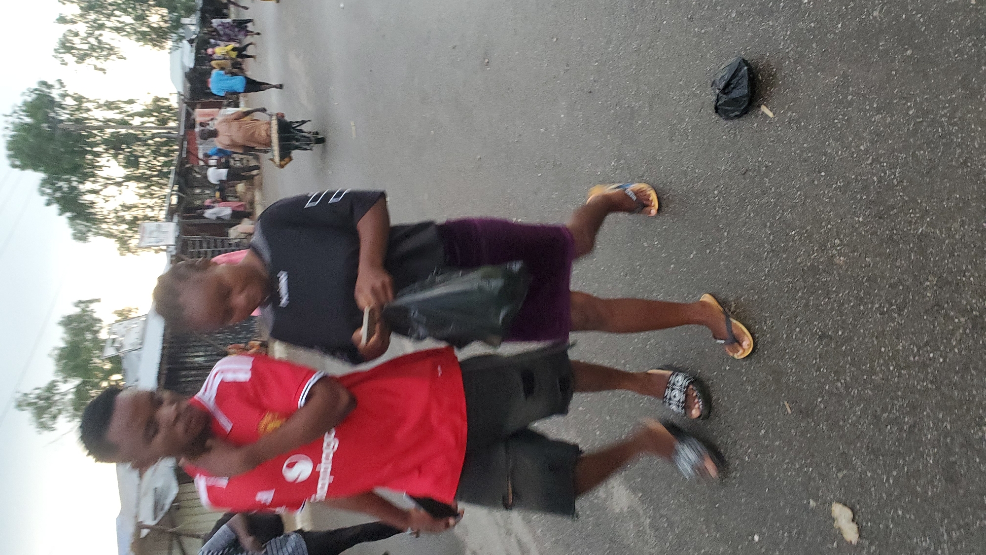 The image depicts a street scene in Jalingo, Nigeria. Two young people, a male and a female, are walking on a paved road. The male is wearing a red t-shirt and dark ripped shorts, with dark sandals. The female is wearing a black t-shirt and a dark purple skirt, with yellow flip-flops. She is carrying a black plastic bag. In the background, there are several small shops or market stalls lining the street, with people milling about. A man with a wheelbarrow loaded with goods is also visible in the background. The sky is overcast.