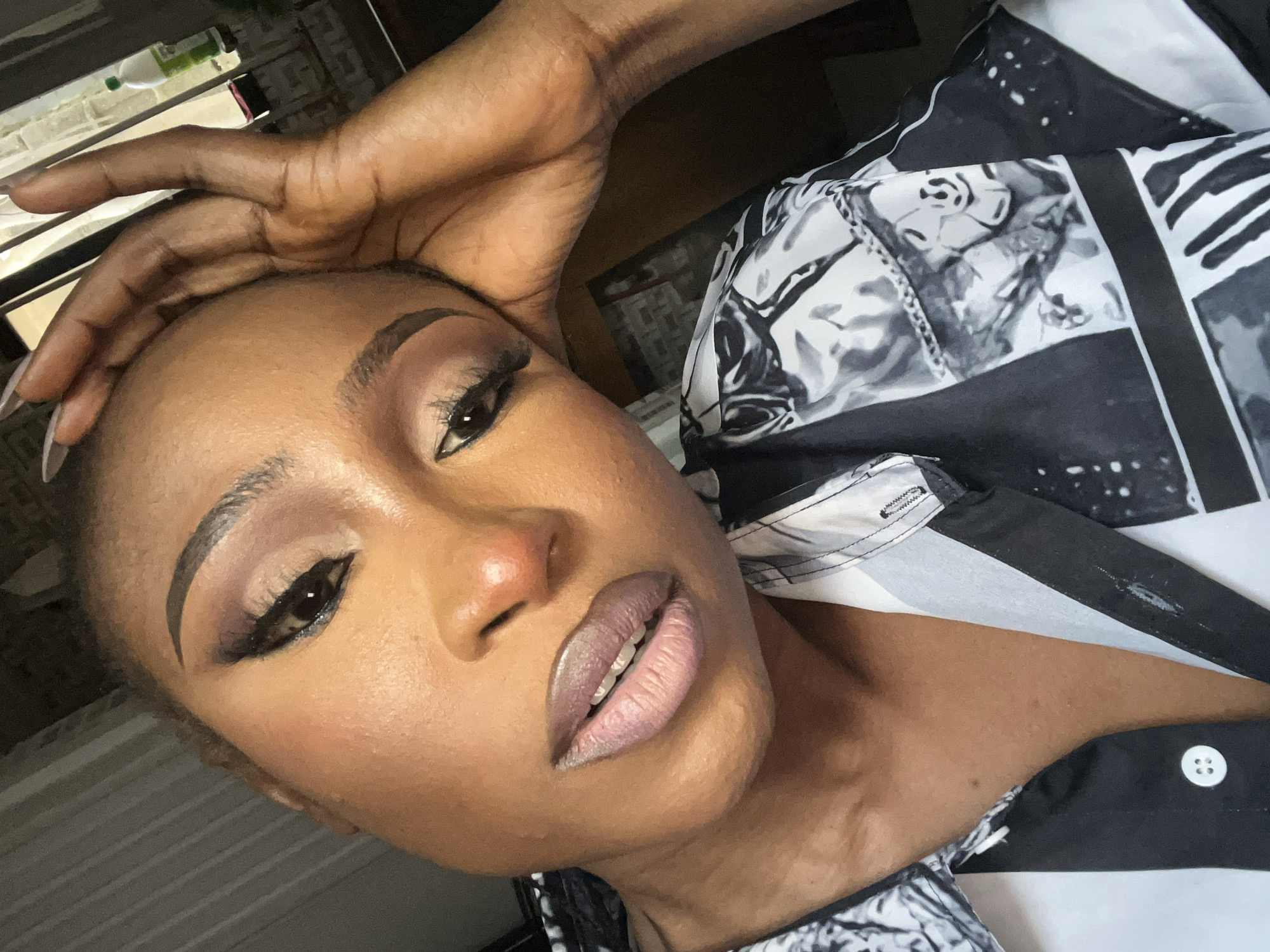 A close-up, overhead shot captures a Black woman with her hand resting on her forehead, looking directly at the camera. She is wearing a black and white patterned shirt. Her makeup includes dramatic winged eyeliner, eyeshadow, and dark lipstick. The background is dimly lit, with vertical blinds visible on the left and a window with a bottle on the right. The image appears to be taken indoors, likely in a residential setting within Abuja, Nigeria.