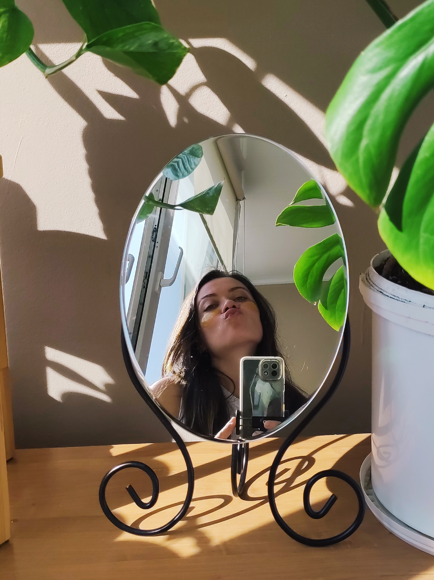 A fair-skinned woman with brown hair is reflected in a round, free-standing mirror on a wooden surface. She is making a kissing face, and there are small, possibly cosmetic, patches under her eyes. She is holding a cell phone with multiple lenses, presumably taking the picture. The phone case has an image on it. Behind the mirror, a window with closed blinds is visible. A green plant, possibly a pothos, is on the left and a larger, potted plant with split leaves is on the right. The background wall is a pale beige. Shadows from the plant are cast on the wall. The location is likely indoors. There is no information in the image to verify it is in Chaikovsky, Russia.