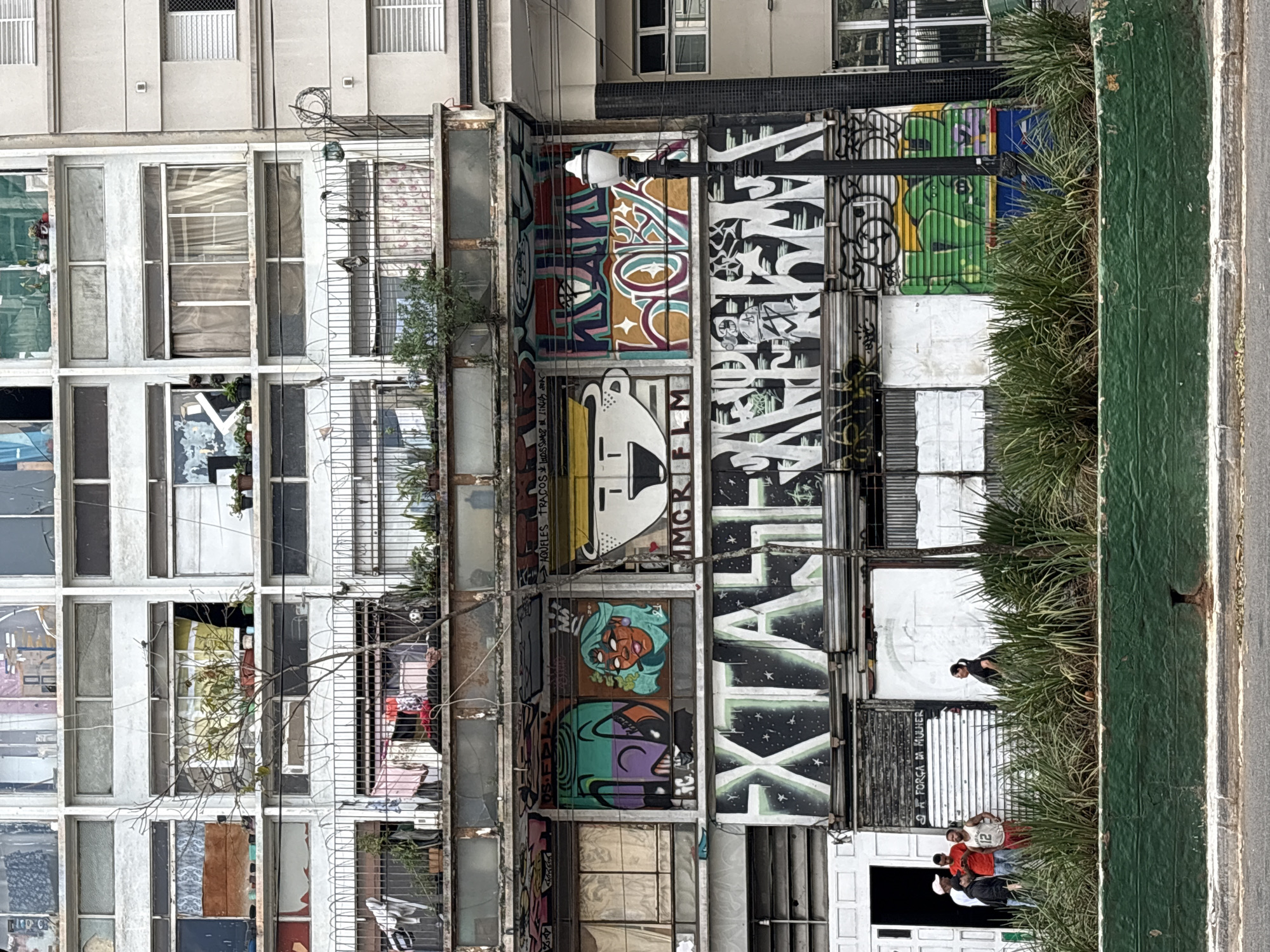 This image depicts a street-level view of a multi-story building in São Paulo, Brazil, heavily adorned with graffiti. The building's facade is weathered and shows signs of neglect, with many windows boarded up or covered with various materials. The lower levels of the building are particularly covered in vibrant graffiti murals, including depictions of stylized faces, text in Portuguese, and abstract designs.
In the foreground, a low, green-painted concrete planter box is filled with dense green foliage. Beyond the planter, a sidewalk and street are visible.
On the ground floor, there is an open doorway leading into a dark interior, and several people are standing outside, some in conversation. Among them, a group of four men, one wearing a red shirt and another a white shirt with the number 12, are visible. A woman is also seen walking away from the group towards the right side of the frame.
A black lamppost with a white fixture stands prominently in front of the building, partially obscuring some of the graffiti. The overall impression is one of urban decay combined with artistic expression.