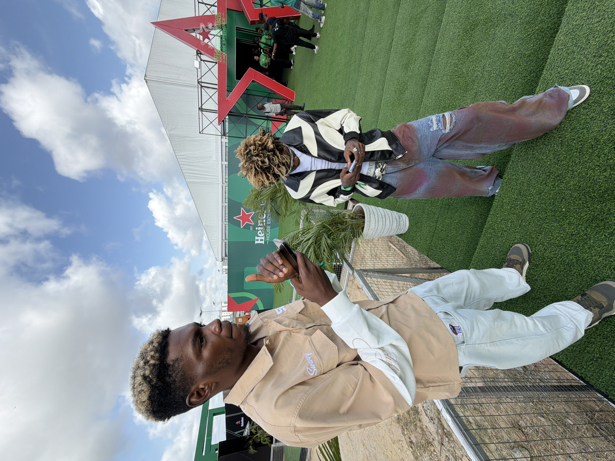 The photograph shows two young men at an outdoor event. The man on the left is wearing a tan jacket with white sleeves and light-colored pants. He holds a smartphone in his hands. The man on the right is wearing a black and white leather jacket over a white shirt and ripped jeans. In the background is a large Heineken sign with a red star, indicating sponsorship. The event appears to be taking place on artificial turf. Given the Heineken branding, the event could be in Lagos, Nigeria, as Heineken is a popular beer brand there and hosts events in the city.