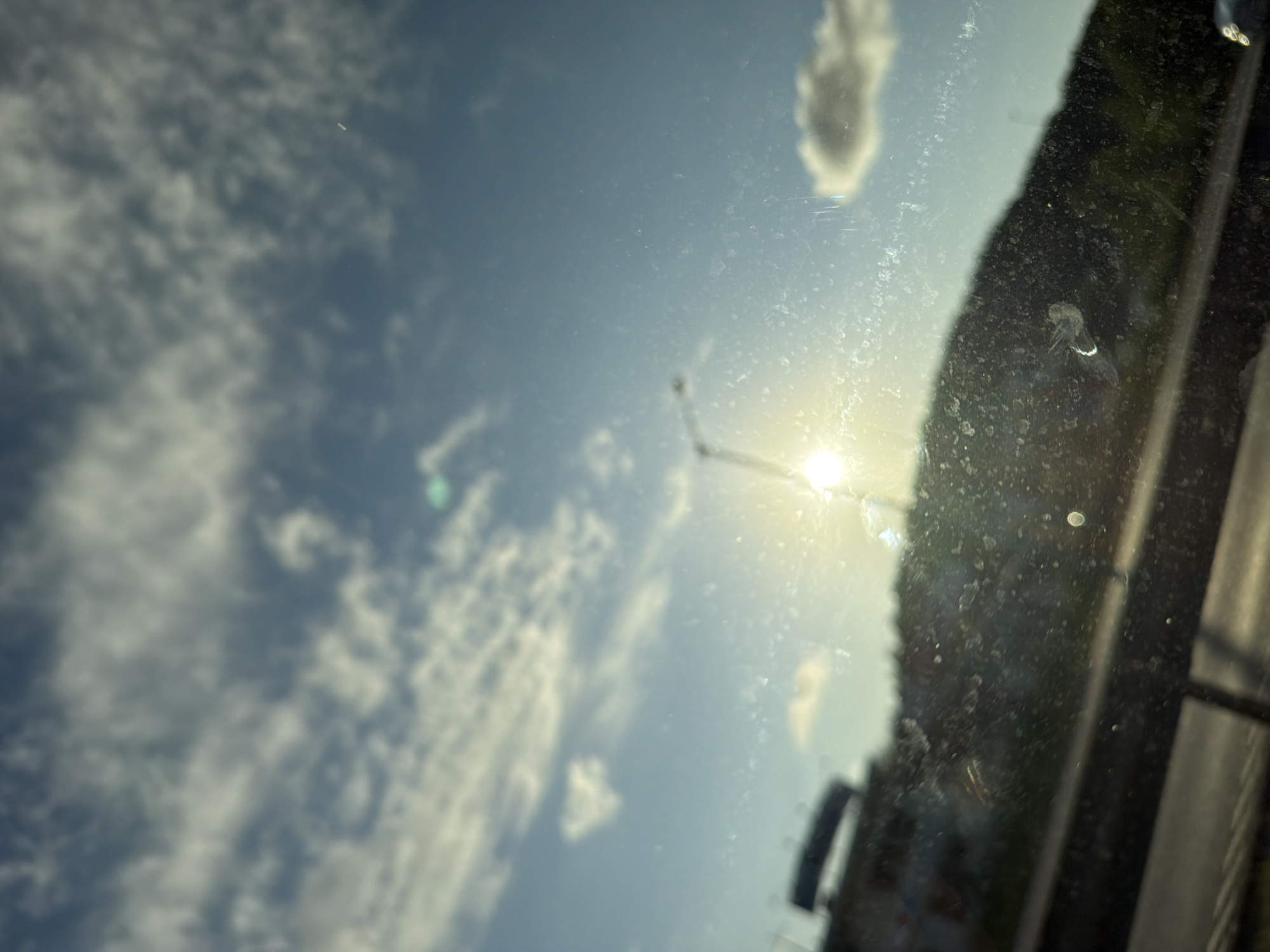 The image is a photograph taken through a dirty windshield, looking out at a bright, sunny sky with scattered clouds. The sun is prominently visible, creating a strong glare and lens flare effect. In the foreground, the windshield has numerous smudges and water spots. Beyond the windshield, a dark, silhouetted hill or mountainside is visible, with some green foliage. To the left, there is a blurred structure that appears to be a building or part of a cityscape, possibly with signage. A street light or pole is also visible on the hill, partially obscured by the sun's glare. The overall impression is of a scenic view from a moving vehicle, likely on a highway, with the sun low in the sky, suggesting either early morning or late afternoon. The location of Mölndal, Sweden, is not discernible from the image itself, as there are no identifying landmarks or contextual clues.