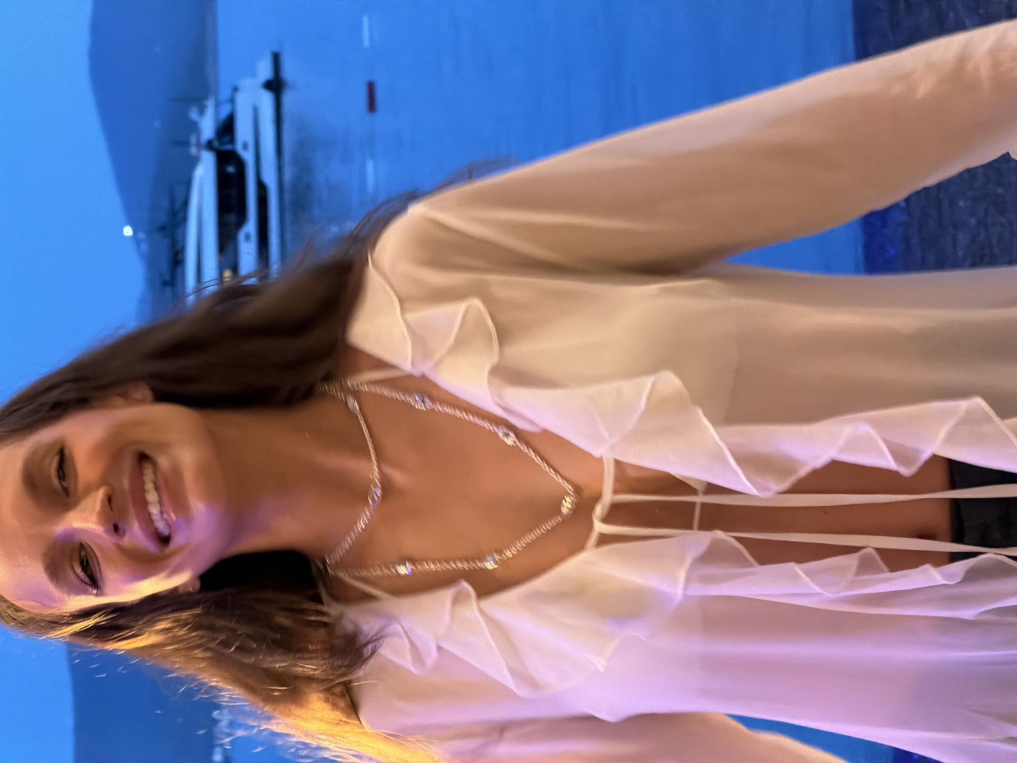 A woman smiles at the camera in Glavaticici, Montenegro. She is wearing a white, frilly top and a silver necklace. In the background, a modern yacht is docked on the blue water. The scene appears to be lit with artificial light.