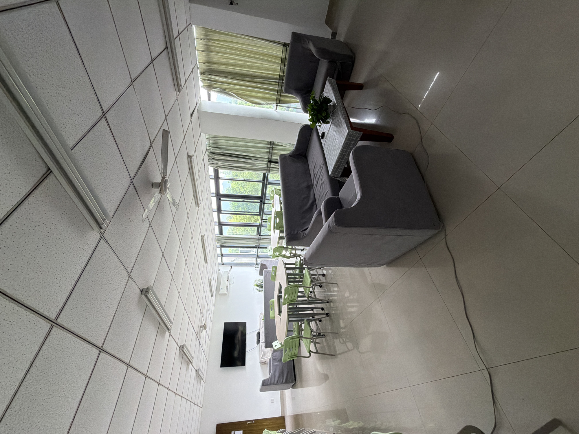 The image depicts a well-lit, modern common area, likely a lounge or break room, with ample natural light filtering in through large windows. The flooring is tiled in a light neutral color, and the ceiling features a standard drop-tile design with fluorescent lighting panels and a ceiling fan. The furniture consists of several grey fabric sofas and armchairs arranged around a low, rectangular coffee table. This coffee table is covered with a checkered cloth and holds a potted plant and a remote control. In the background, there are round and rectangular tables accompanied by bright green chairs, suggesting a dining or meeting space. A large flat-screen television is mounted on a white wall, and a dark-colored door is visible to the left. Outside the windows, lush green trees and foliage are visible, indicating a location with a natural environment. The overall impression is one of a clean, organized, and comfortable space designed for relaxation or informal gatherings. The image does not contain any individuals, and therefore no actions or interactions are observable. The style of the room suggests it is located within an institutional setting, possibly a school, dormitory, or office complex, in the city of Jiangjin, China.