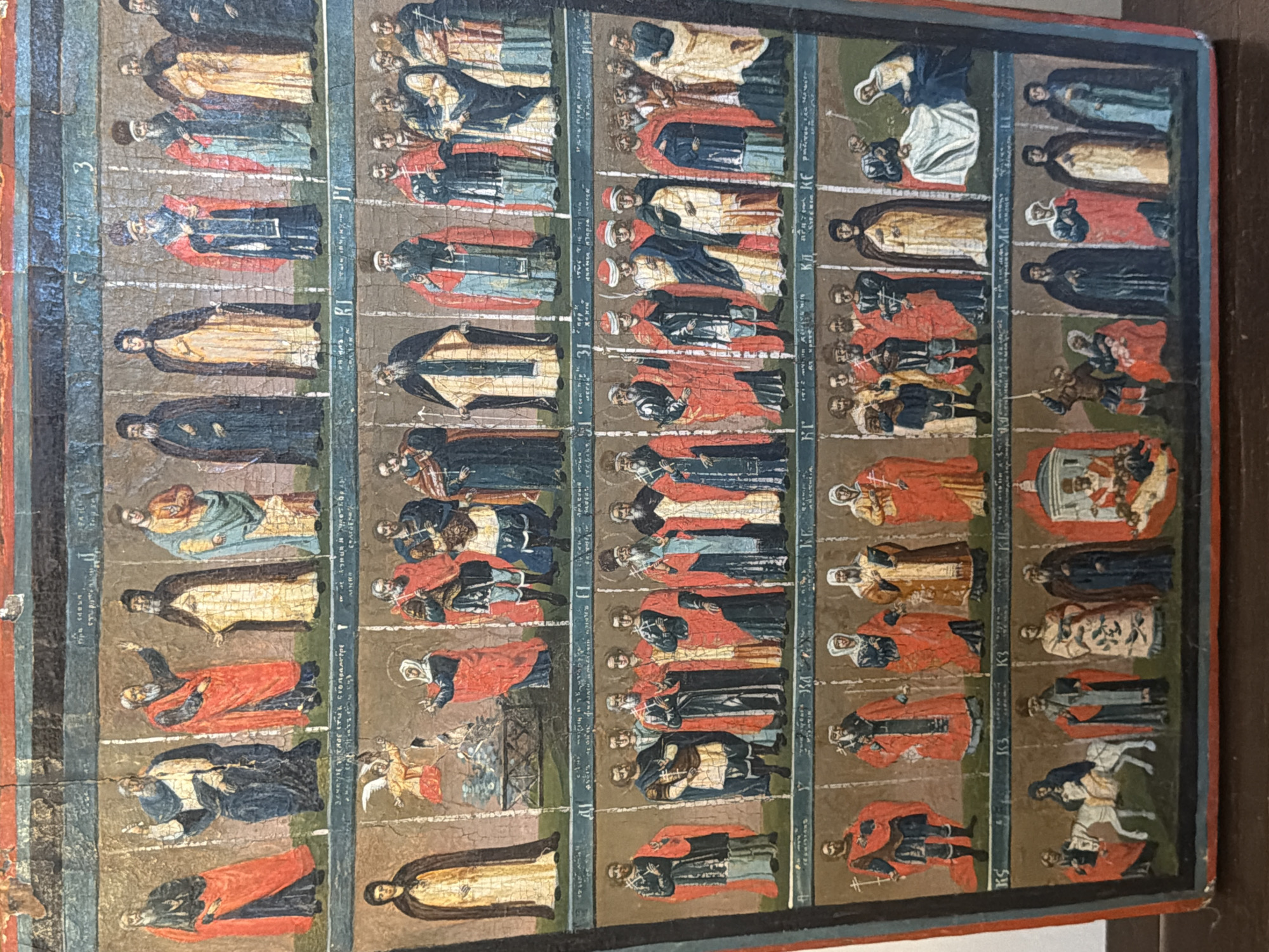 The image is a painting of religious figures, likely an icon, arranged in rows. The figures are dressed in robes of various colors, including red, blue, yellow, and white. Many of the figures have beards and are depicted with halos, indicating they are saints or religious leaders. Some figures hold religious symbols such as crosses.
The painting is divided into horizontal bands, with each band containing multiple figures. The figures are depicted standing in front of plain backgrounds, with some subtle architectural elements or landscapes in the background of some scenes. The style of painting appears to be traditional, with flat figures and a lack of deep perspective. There are also small inscriptions written in an unfamiliar script above and between some of the figures. The overall condition of the painting shows signs of age and wear, with cracking and flaking paint visible in several areas.
There is no indication in the image or the provided crops that this painting is located in Ko Samui District, Thailand. The style of the artwork and the script suggest a Christian religious context, likely Eastern Orthodox.