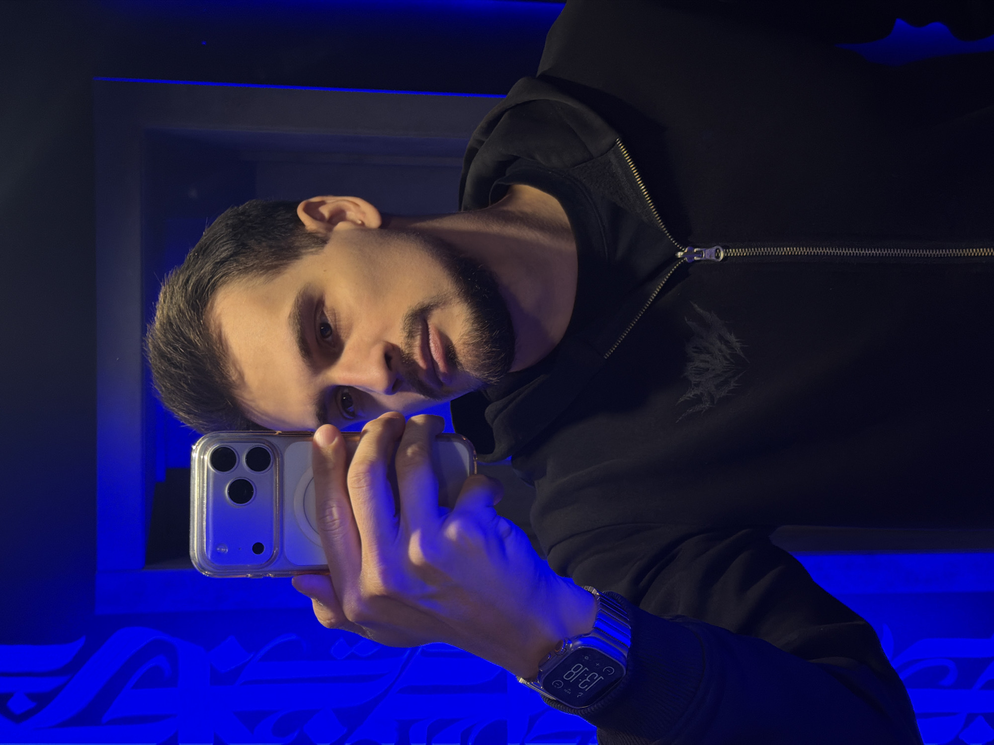 A man is taking a selfie in what appears to be a location in Krasnoyarsk, Russia. The man has short brown hair, light skin, and a short beard. He is wearing a black hooded sweatshirt and a stainless-steel watch on his left wrist. He is holding a smartphone with a clear case in his left hand. The phone is positioned to capture a selfie of the man. The background is dark with blue neon lighting. The room appears to have decorative designs illuminated by the blue light. A framed area with dark gray borders is visible above the man.