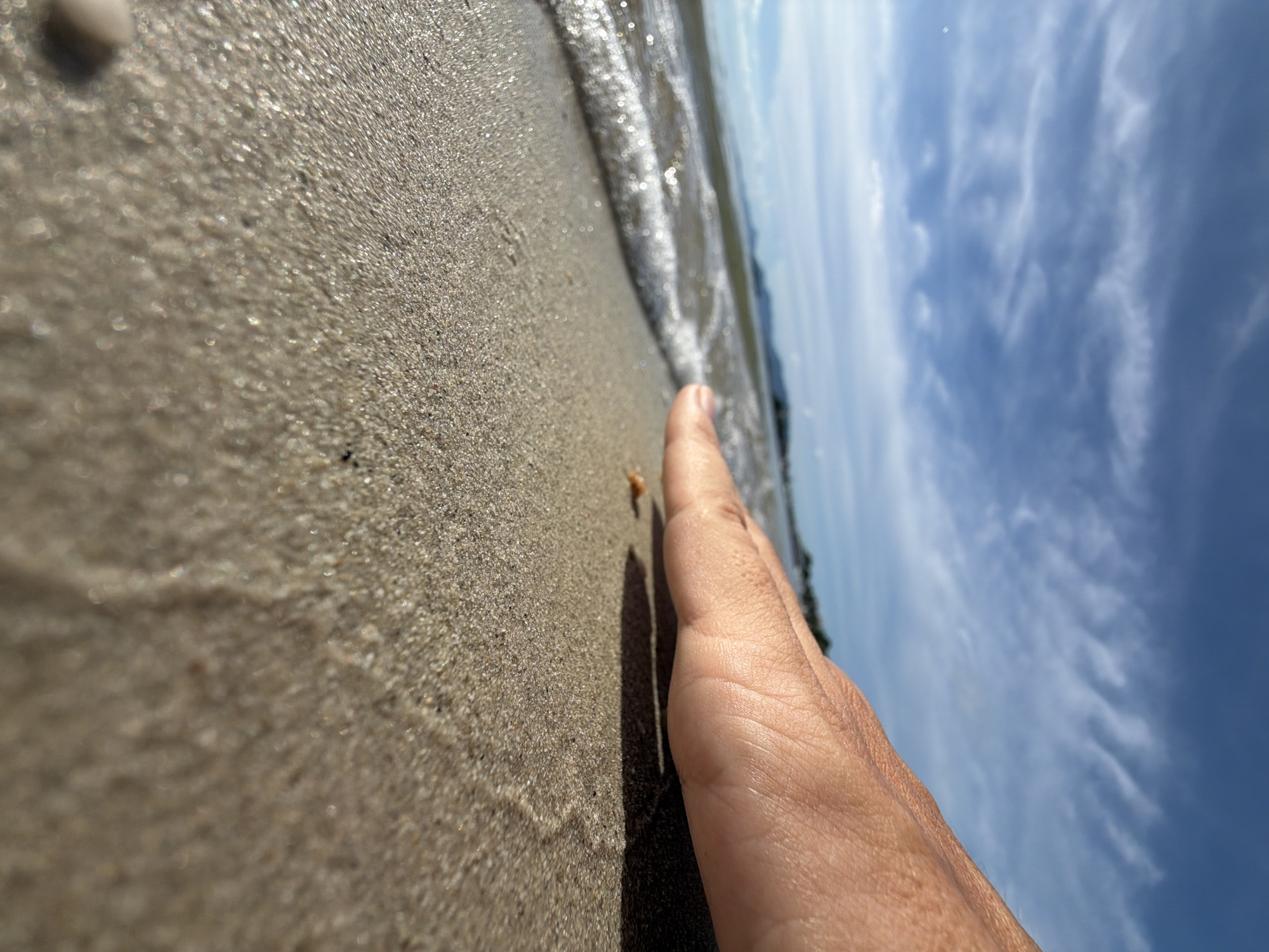 The media file shows a close-up, low-angle shot of a sandy beach with the ocean and sky in the background. A person's hand, specifically the palm and fingers, is in the foreground, extended towards the water. The sand is light brown and appears wet near the shoreline, with small ripples from the receding waves. The ocean is calm with a gentle wave breaking. The sky is a clear blue with wispy white clouds, suggesting a sunny day. The lighting indicates it is likely daytime, possibly late morning or afternoon, given the bright sunlight and clear visibility. The scene evokes a sense of relaxation and enjoyment of a beach environment. The location is contextually identified as Baan Bo Phut, Thailand. There is no visible text in the media file.
