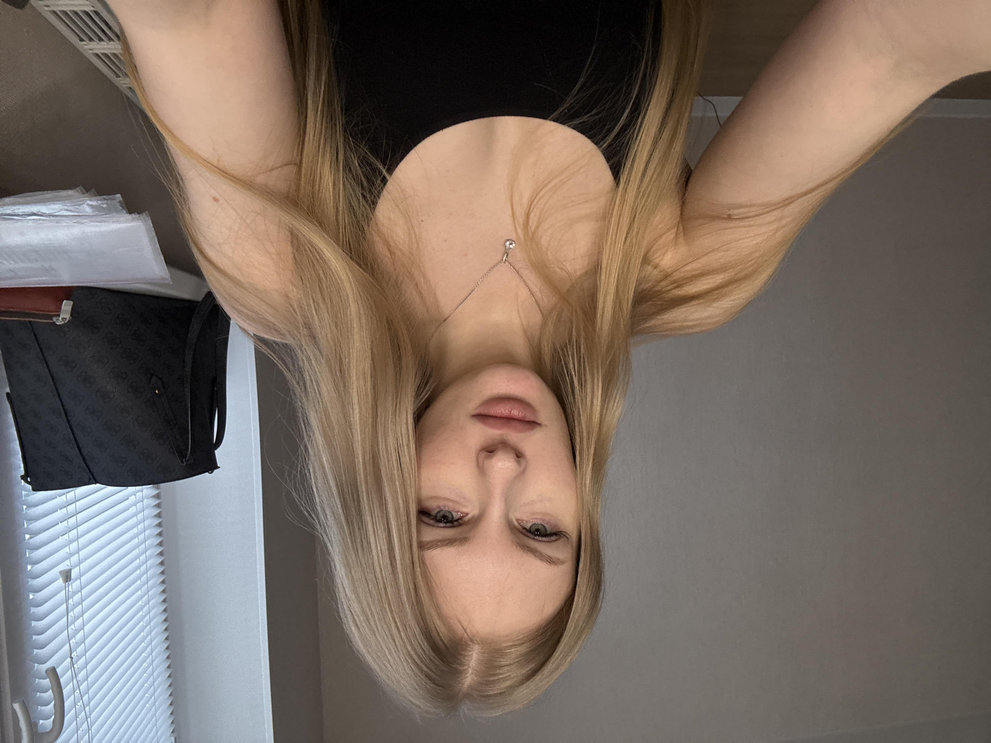 An inverted close-up portrait features a young woman with long, straight, blonde hair, parted in the middle. Her head is positioned towards the bottom of the frame, with her hair flowing upwards and to the sides. She has fair skin, full lips slightly parted, and light-colored eyes that gaze directly at the viewer with a neutral expression. Her eyebrows are well-defined, and visible eyelashes frame her eyes.
She wears a simple black top, and a delicate silver-colored necklace with a small, clear gemstone pendant rests on her visible décolletage. Her arms are visible, extending upwards and outwards from her body. A small, dark mole is noticeable on the inner aspect of her right arm (which appears on the left side of the image).
The background elements suggest an indoor setting. To the left side of the frame (behind her right arm), a patterned black bag with a repeating 'G' motif (resembling a Guess logo) is visible, hanging or placed near a window. Below the bag, a white window blind with horizontal slats is prominent, alongside what appear to be white plastic bags or packaging. Above the bags and near the top left corner, a white vent is mounted on a wall. The right side of the background (behind her left arm) consists of a plain, light grey wall.
The lighting is soft and even, suggesting indoor conditions, possibly with some natural light filtering through the window during the daytime. The image captures the woman in a contemplative or posed manner, likely a self-portrait taken from an overhead perspective. The location context for this image is Perm, Russia.