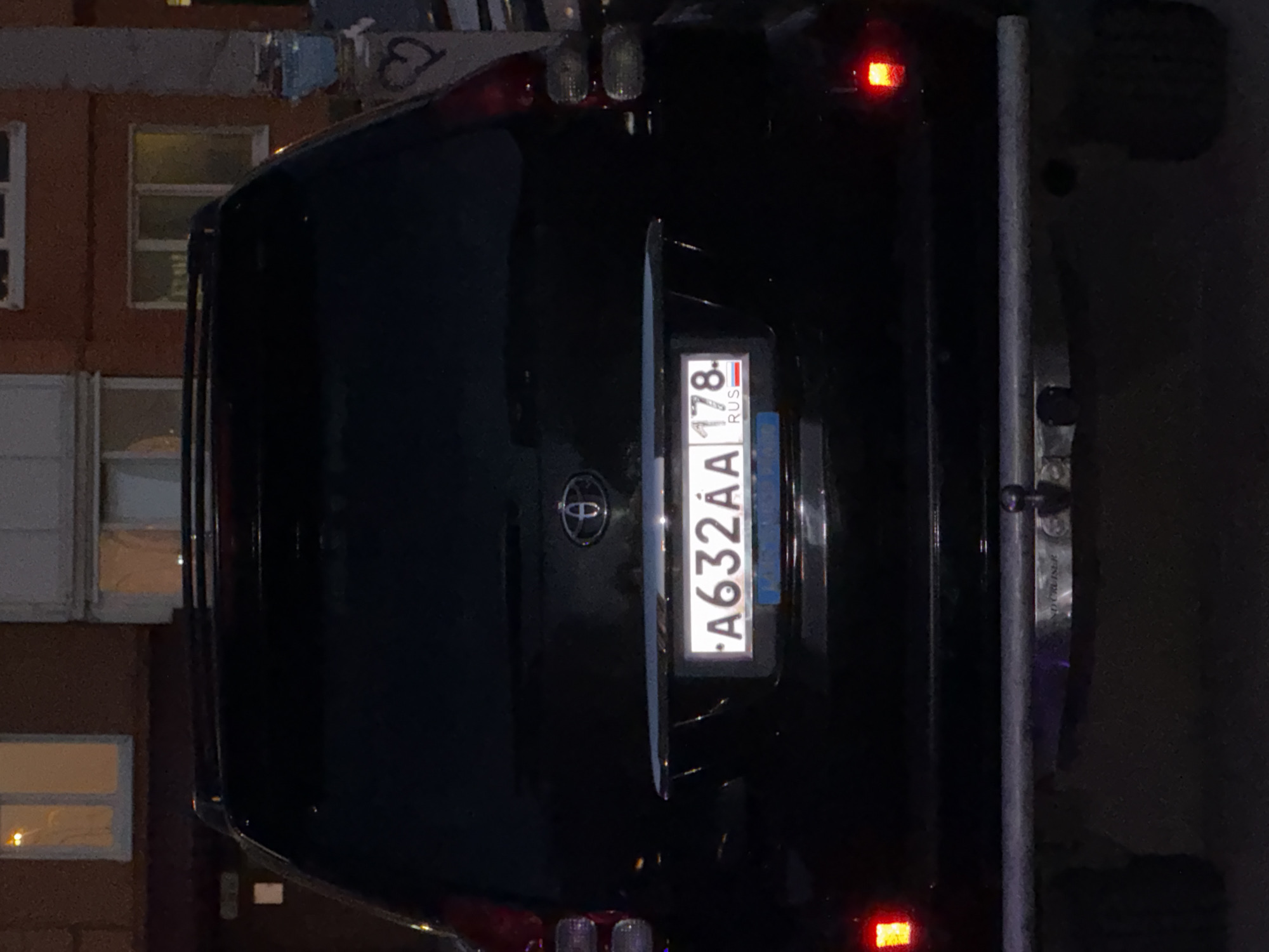The image shows the rear of a black Toyota Land Cruiser Prado SUV parked at night. The license plate reads "A632AA 178 RUS". The vehicle is parked on a paved surface, possibly a street or parking lot, in front of a building with brickwork and windows. There are no people visible in the image. The lighting suggests it is nighttime, with artificial lights from the building and the vehicle's taillights illuminating the scene. The environment appears to be an urban setting, likely in Saint Petersburg, Russia, as indicated by the license plate's region code "178".