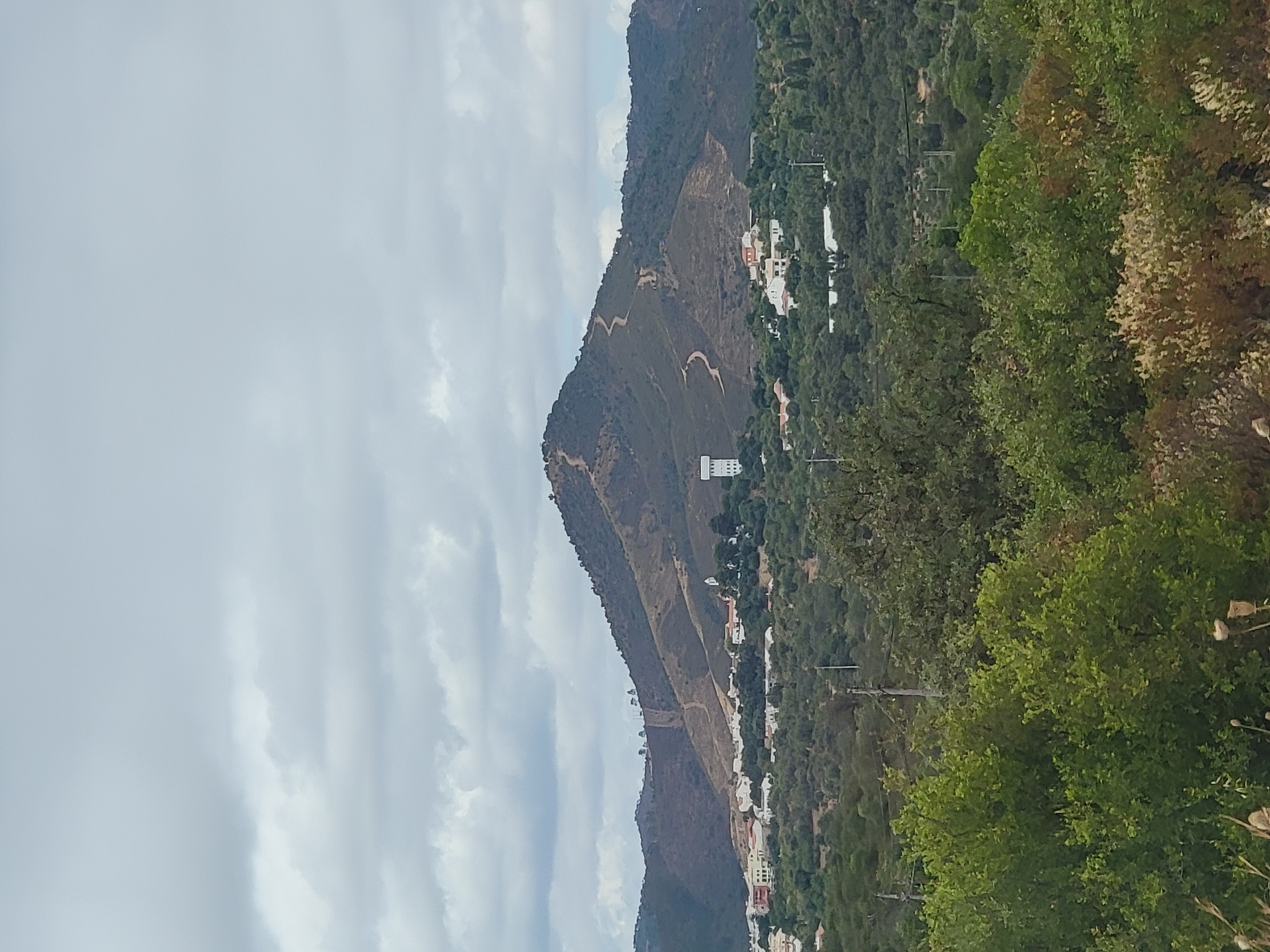 The image depicts a scenic view of the town of Loulé, Portugal, characterized by rolling hills and a vast expanse of greenery. Dominating the landscape is a prominent mountain with a winding path leading up its slopes, likely a trail for hiking or access. Perched on the mountainside is a tall, white water tower, a recognizable landmark. Below the mountain, the town of Loulé sprawls across the landscape, featuring clusters of white-washed buildings with red-tiled roofs. The foreground is lush with trees and vegetation, suggesting a rural or semi-rural setting. The sky is overcast with scattered clouds, contributing to a soft, diffused light. The image captures a tranquil and picturesque aspect of Loulé's surroundings.