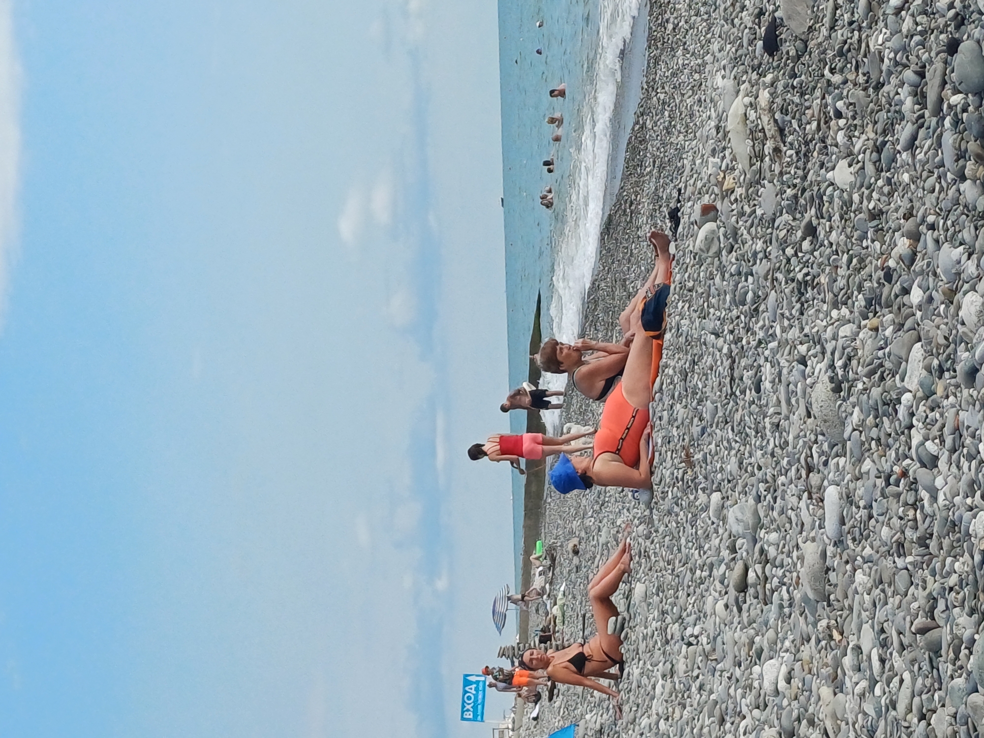 This is a vertical, eye-level, medium shot of a crowded, pebbly beach in Sochi, Russia, on a bright, sunny day. The sky is a clear, pale blue with some scattered white clouds. The sea is a deep blue and waves are gently rolling onto the shore. Numerous people are visible on the beach and in the water, engaged in various activities.
In the foreground, a woman in a black bikini is lying on the pebbles, looking towards the camera. Behind her, a woman in an orange swimsuit and a blue swimming cap is also lying down, looking to her right. Further back, a woman in a red top and pink shorts stands, facing away from the camera. In the water, several people are swimming and wading. A striped umbrella provides shade for some beachgoers. The beach is covered in smooth, grey pebbles.
A blue sign with white text is visible on the left side of the image, reading "ВХОД," which translates to "Entrance" in Russian. The overall atmosphere appears relaxed and summery, with people enjoying a day at the beach. The lighting suggests it is daytime, likely late morning or early afternoon.