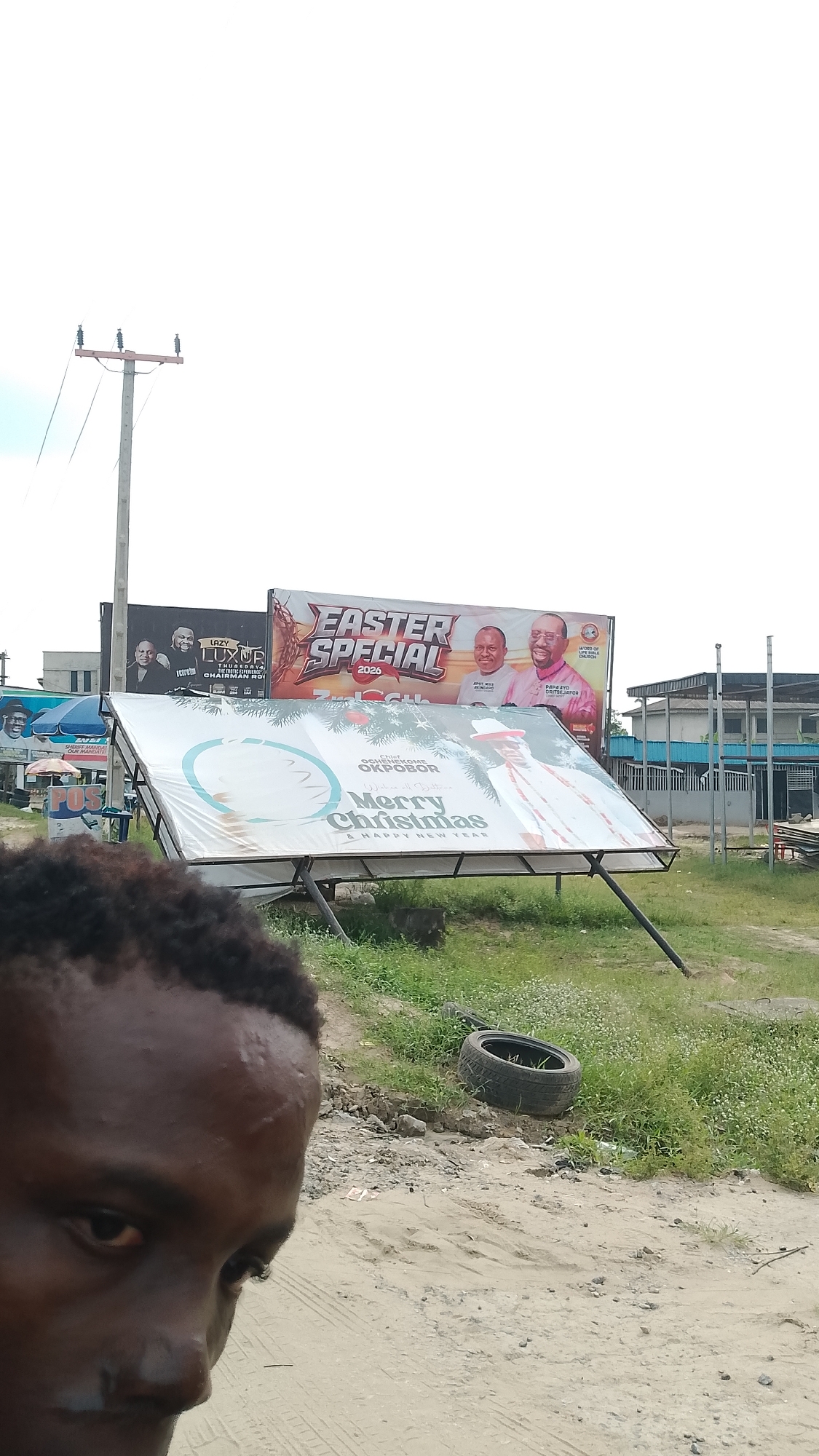 The image shows a partial view of a dark-skinned person in the foreground, with multiple billboards visible in the background. The billboards are displaying various advertisements, including "Easter Special 2026" and "Merry Christmas & Happy New Year." One billboard appears to have fallen or is significantly tilted. The location is indicated by the "Ochenekome Okpobor" billboard which indicates a Warri, Nigeria, location. A tire is discarded on the ground near the fallen billboard. A utility pole is visible in the background, and the general environment suggests a roadside scene.