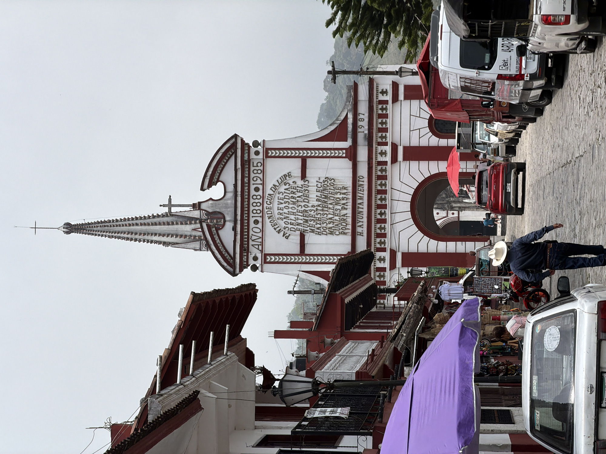 A photograph depicts a vertical view of a street scene in Cuetzalan, Mexico. The architecture is characterized by buildings painted in red and white. One building is topped with a tall, ornate spire and has an inscription that reads "PANTEONDE GUA DALUPE" and "H. AYUNTAMIENTO." The years "1888-1985" and "1987" are also visible on the building's facade.
The street is lined with parked cars and trucks. A man wearing a hat is captured walking alongside the vehicles, facing towards the building. The scene includes various commercial elements, such as a purple awning or tarpaulin and stalls with baskets and merchandise. The sky appears overcast and white.