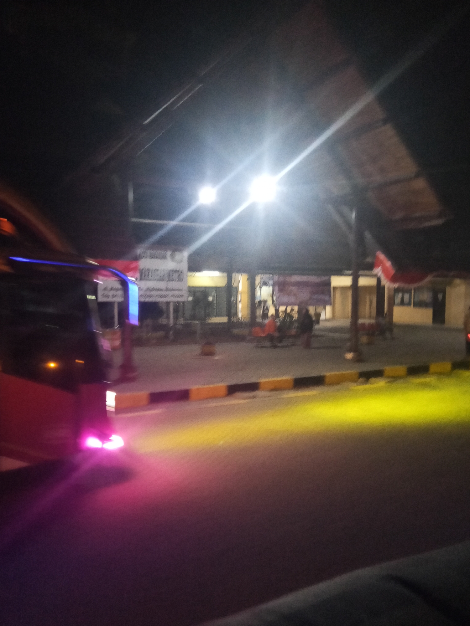 The image shows a bus at a bus terminal at night. The bus is red and has blue lights along the top. The bus terminal appears to be an open-air structure with a roof supported by pillars. There are signs and some people visible in the background. The ground is paved and there are yellow and black striped barriers along the edge of the road. The lighting suggests it is nighttime, with artificial lights illuminating the scene. The image is somewhat blurry and taken from a low angle, possibly from inside another vehicle. The presence of the bus, terminal structure, and signage indicates a transportation hub within the city of Makassar, Indonesia.