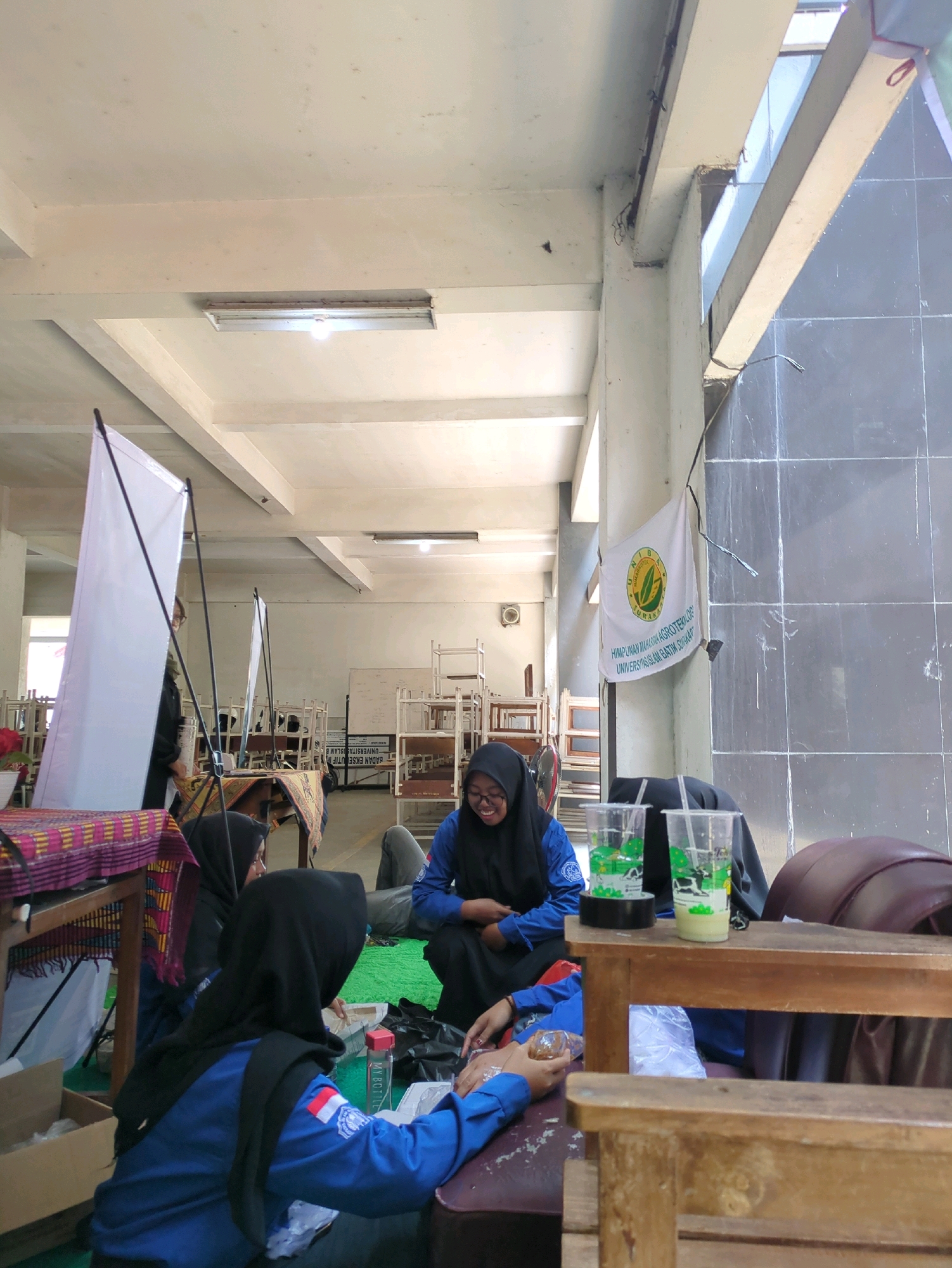 The image depicts an indoor scene, possibly a community hall or a large room, in Surakarta, Indonesia. Several young women, dressed in blue shirts and black headscarves, are gathered. One woman in the foreground, facing away from the camera, wears a blue uniform shirt with a white and red Indonesian flag patch on her left sleeve. She is seated and appears to be interacting with a small, clear container. Next to her, another woman in a similar blue uniform and headscarf is seated and smiling. She is holding a small, clear plastic cup containing a green liquid and a straw. In the background, a white banner with a green logo and text in Indonesian is hung on a wall. The room has a high ceiling with exposed beams and fluorescent lighting. Stacks of chairs and what appear to be display shelves are visible in the background, suggesting a functional space that might be used for events or exhibitions. The overall impression is a casual gathering, possibly related to a student organization given the university banner.